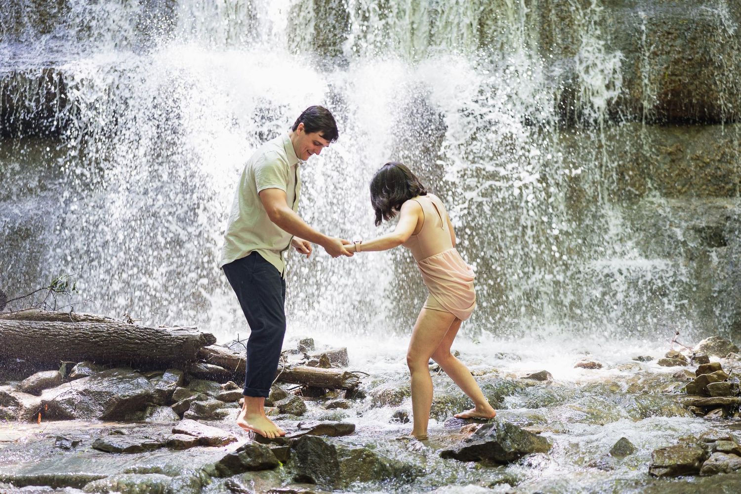 Playful couple holds hands while exploring rocks near a waterfall.