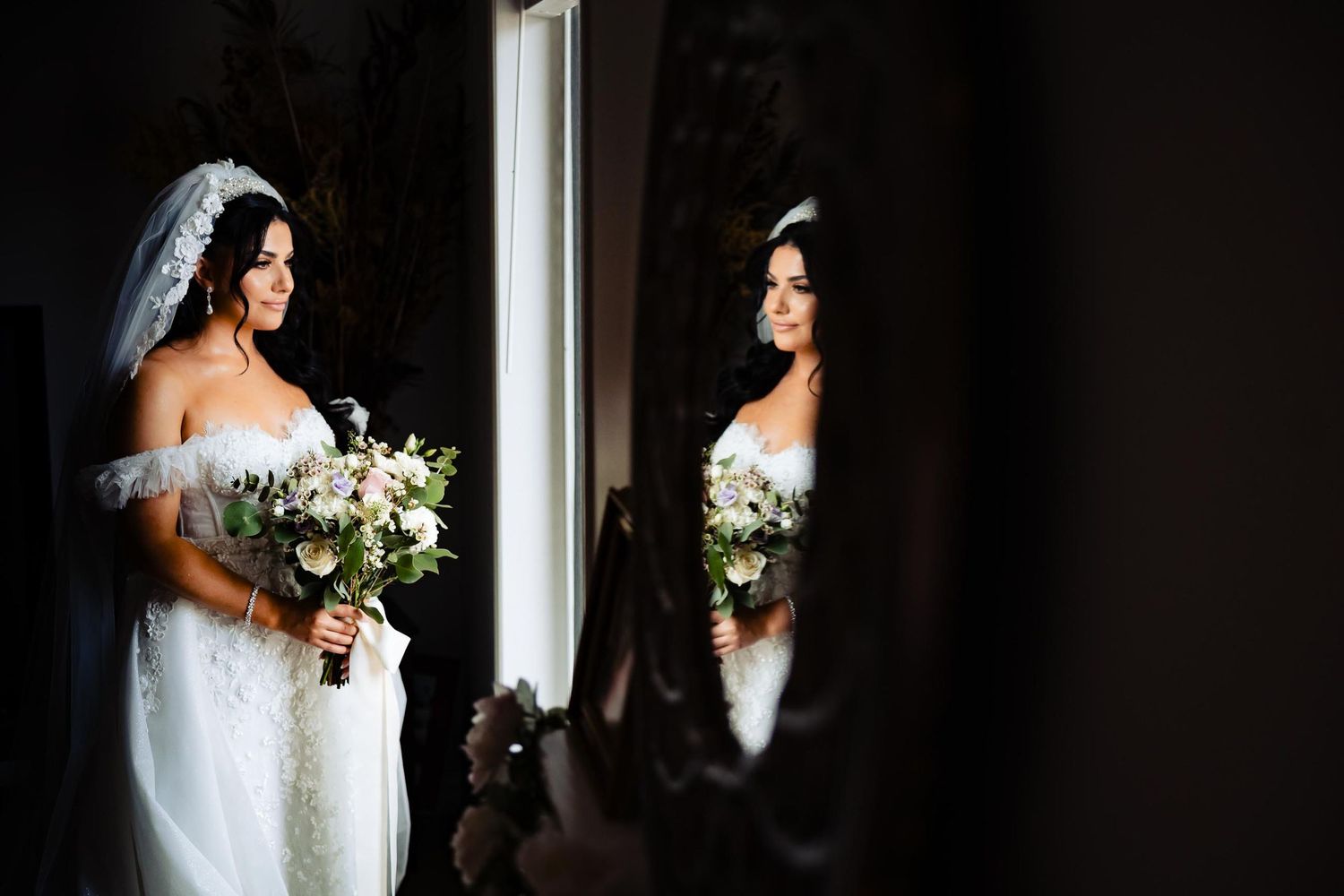 Bride in white dress holding bouquet while looking in reflection of window.