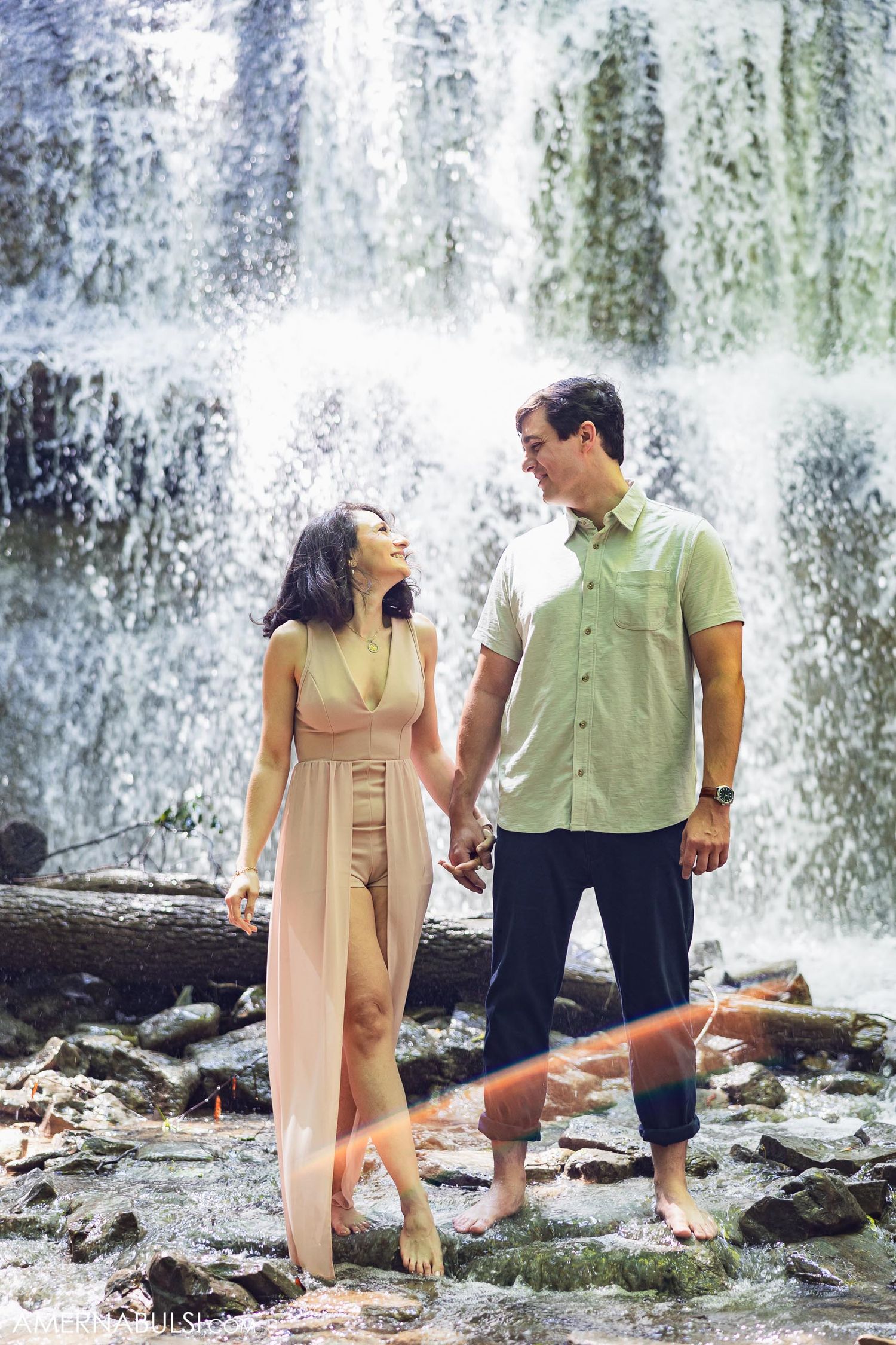 A romantic couple stands barefoot on rocks near a cascading waterfall wearing coordinated casual outfits.