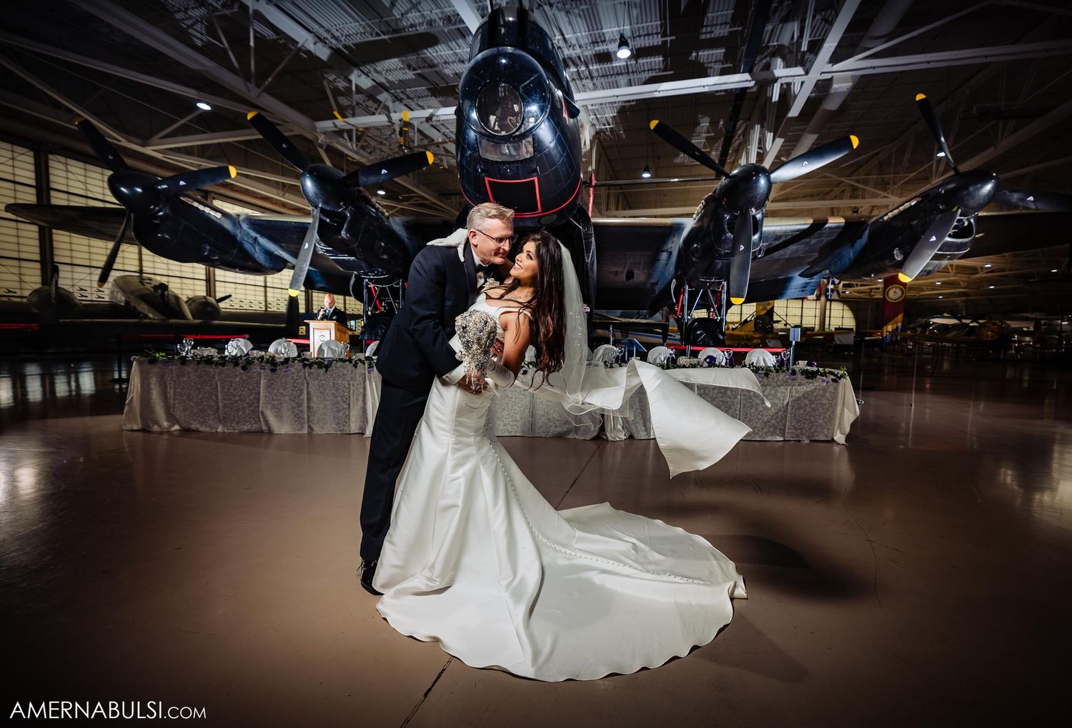 A romantic wedding photo captured beneath a vintage aircraft in a museum setting.