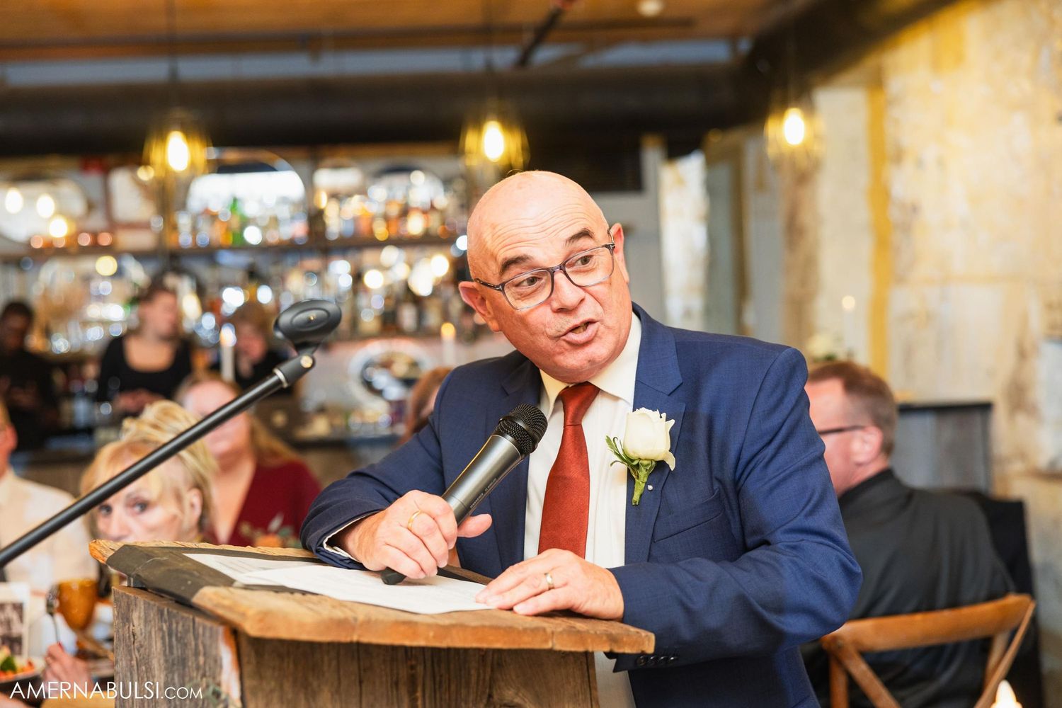 A speaker in a blue suit and glasses delivers remarks at a podium during an event.