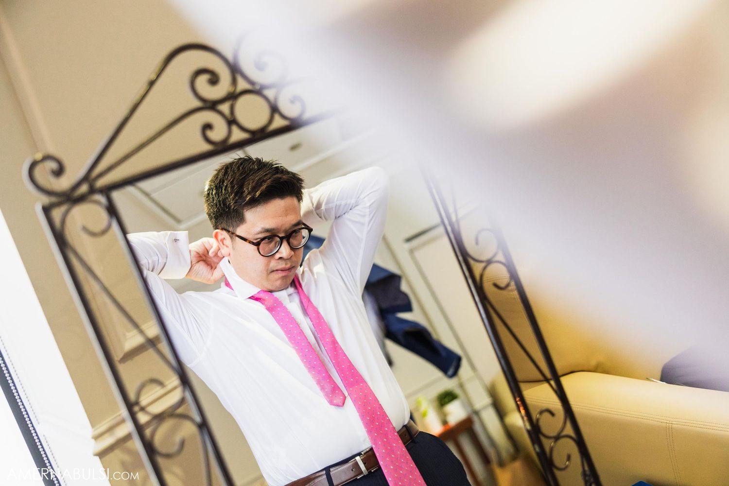 Person adjusting pink tie while looking in ornate mirror during wedding preparations.