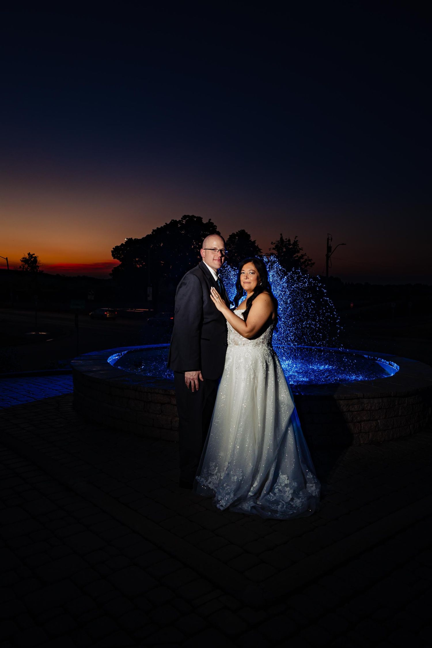 Couple in wedding attire pose by illuminated fountain at sunset.