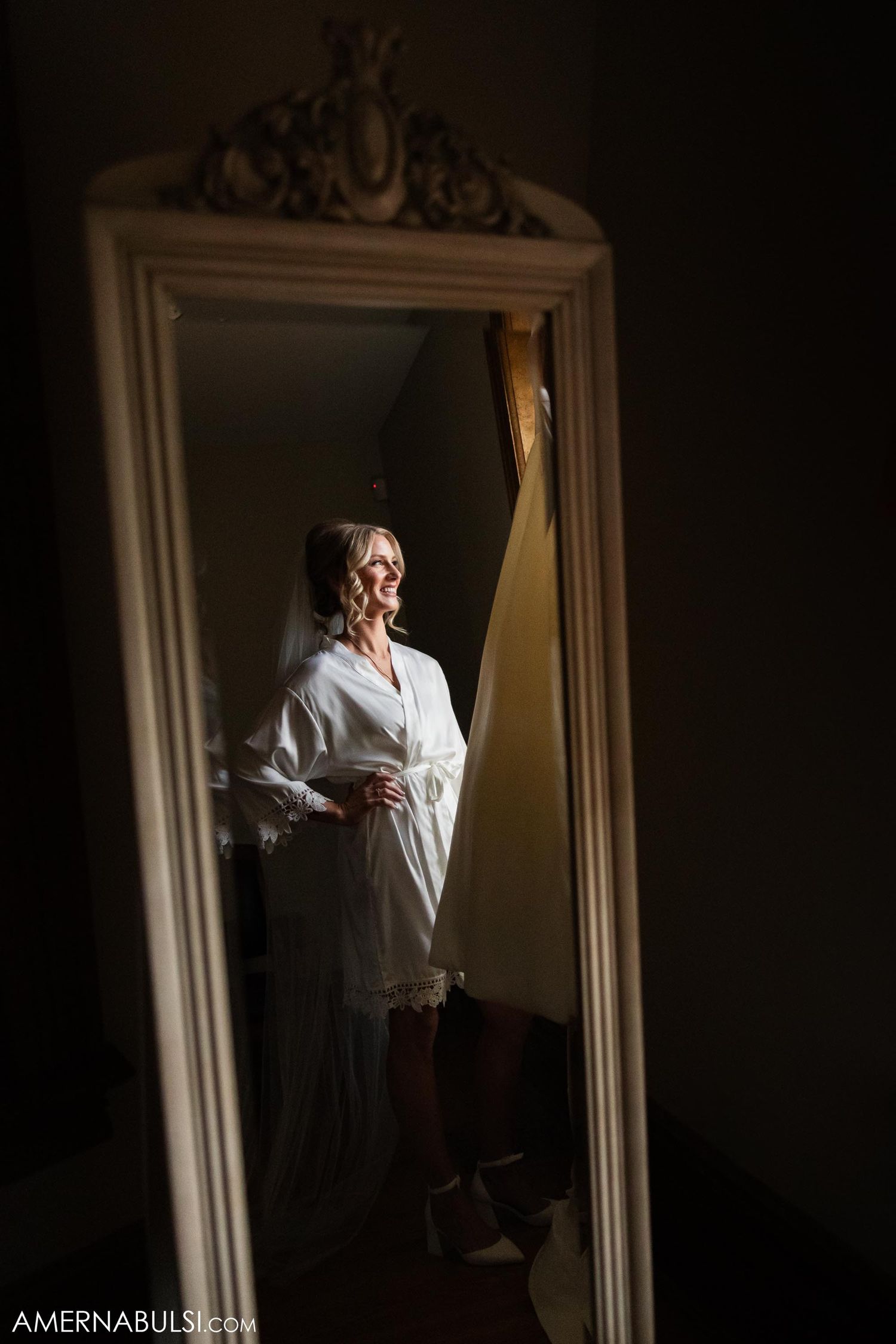 Artistic reflection of bride in white robe through ornate vintage mirror in dramatic lighting.