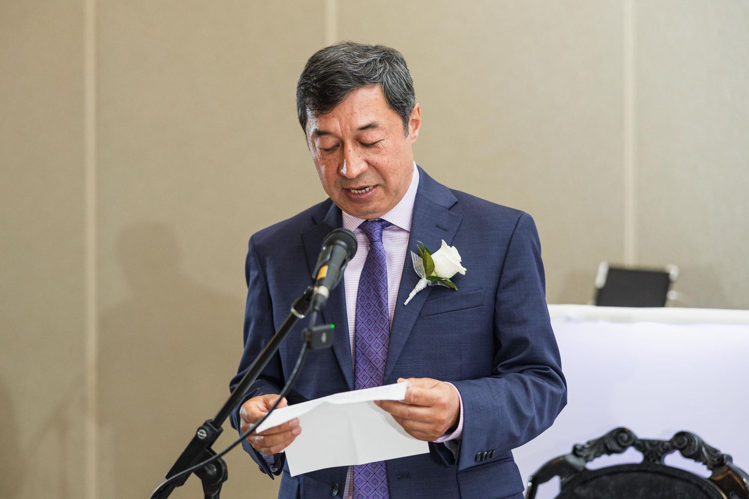 A speaker in a navy suit and purple tie delivers a speech at a formal event with a boutonniere.