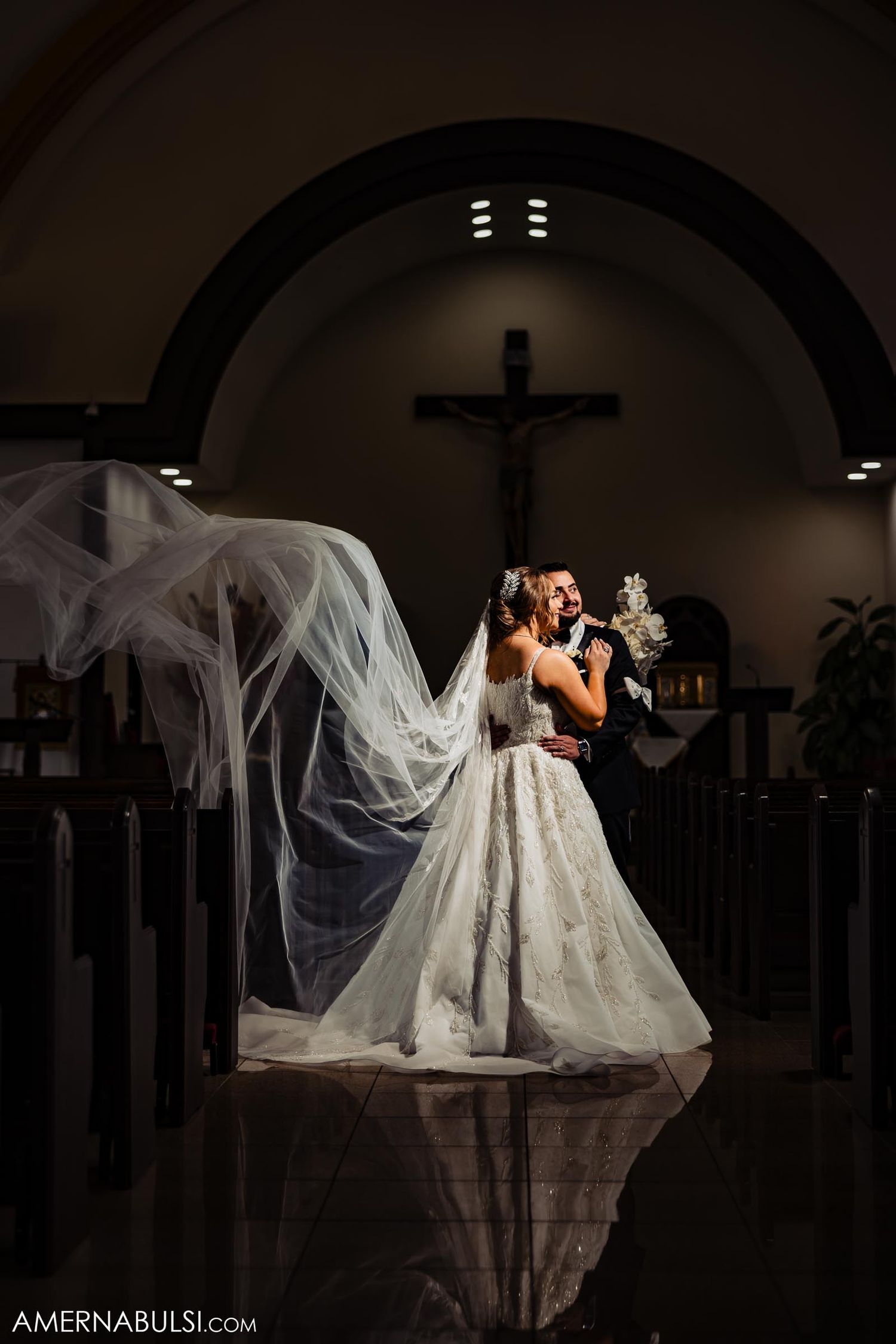 Dramatic backlit wedding photograph in church with flowing veil and crucifix in background.