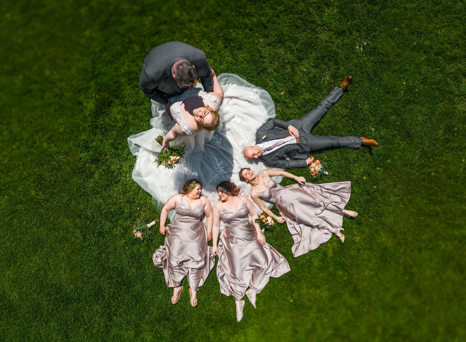 Aerial view of wedding party lying in circle formation on green grass.