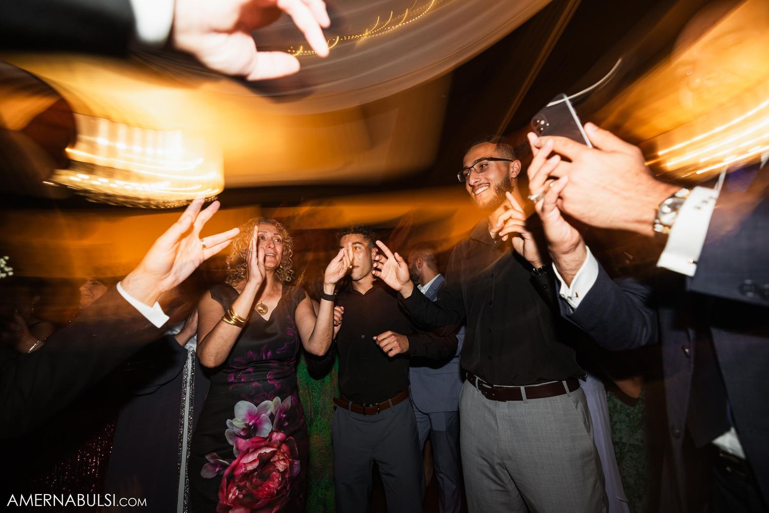 Wedding guests dancing and celebrating energetically under warm lighting at an evening reception.