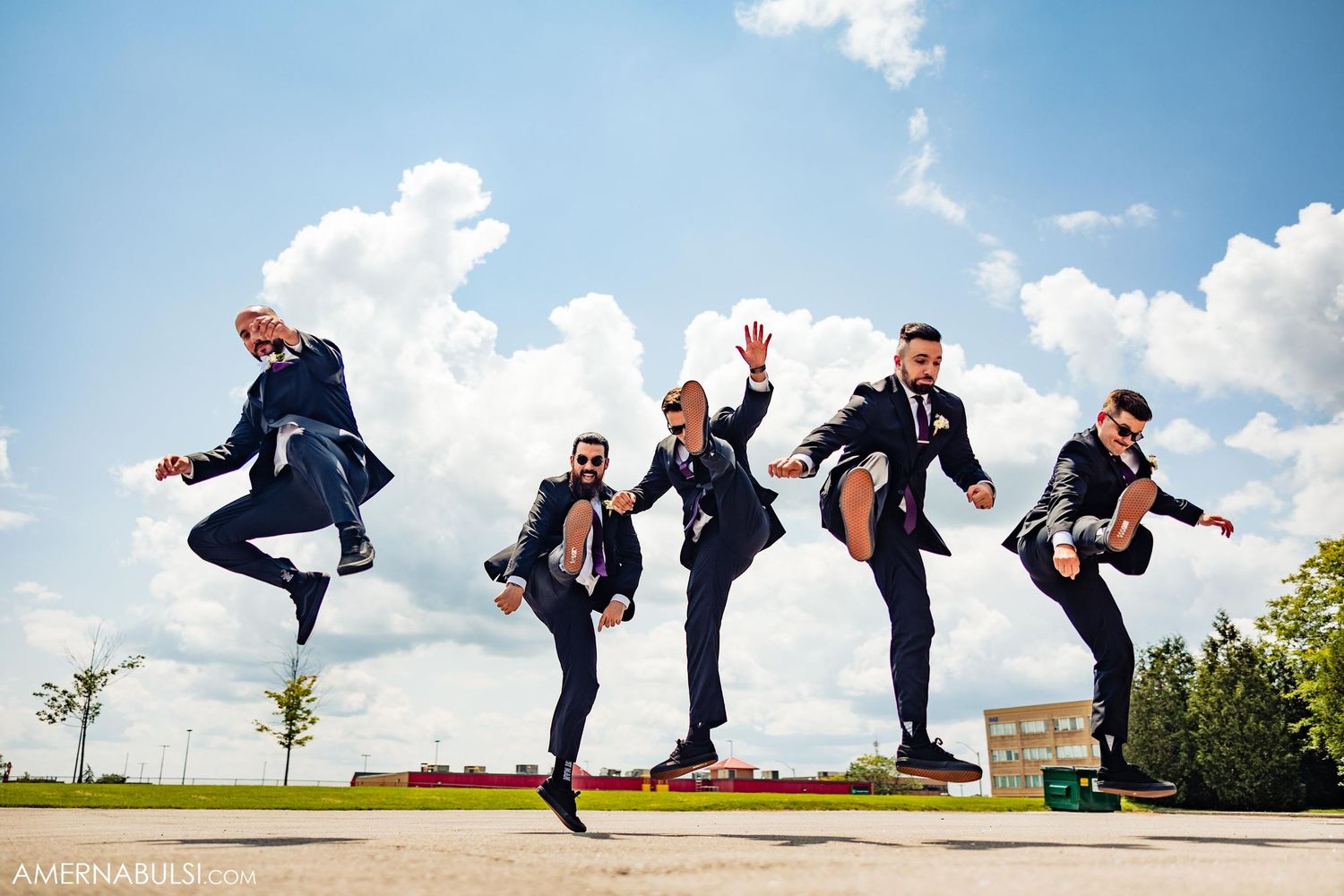 Groomsmen jumping in sync against blue sky in navy suits.