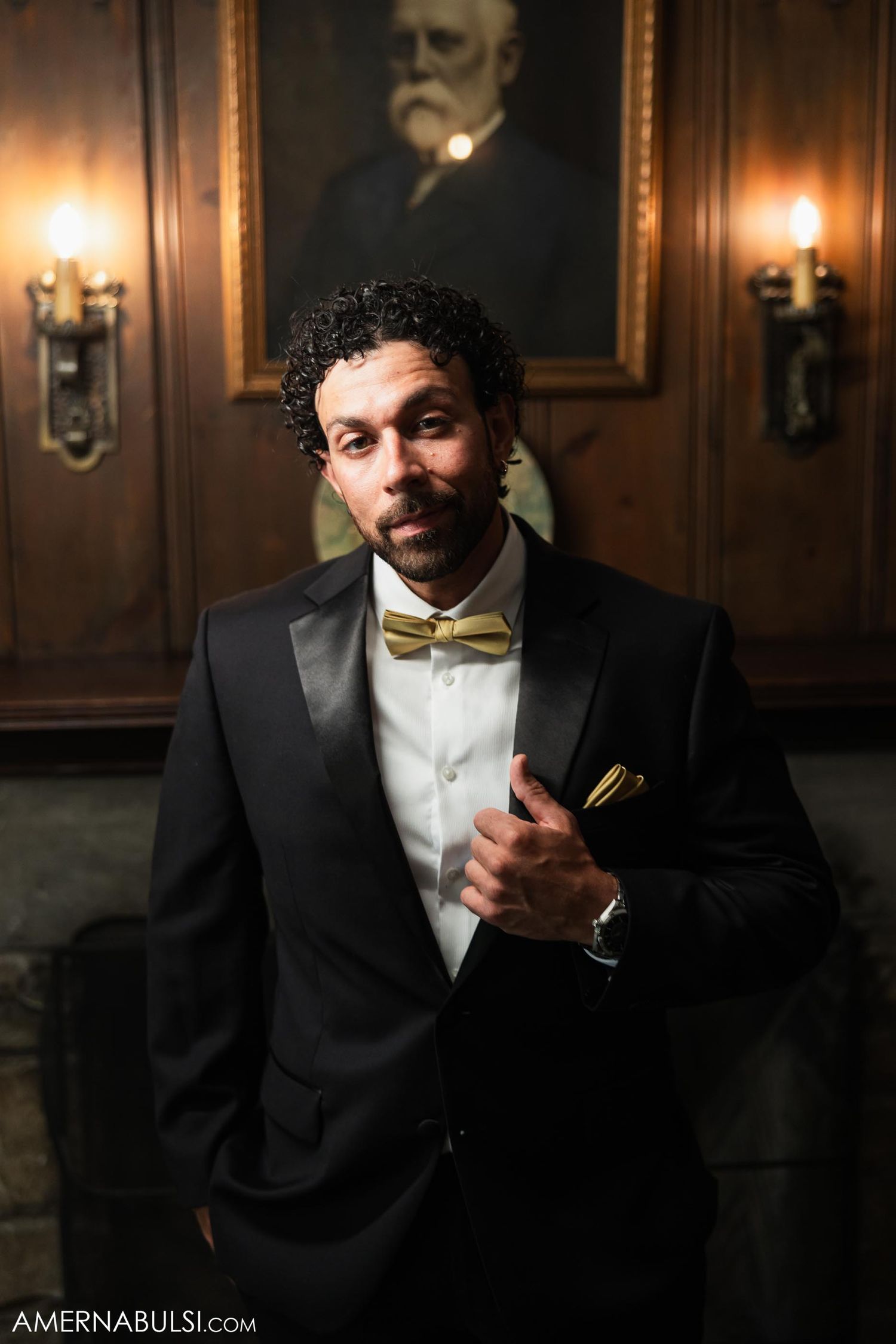 Person in formal tuxedo and gold bow tie poses beneath an antique portrait.