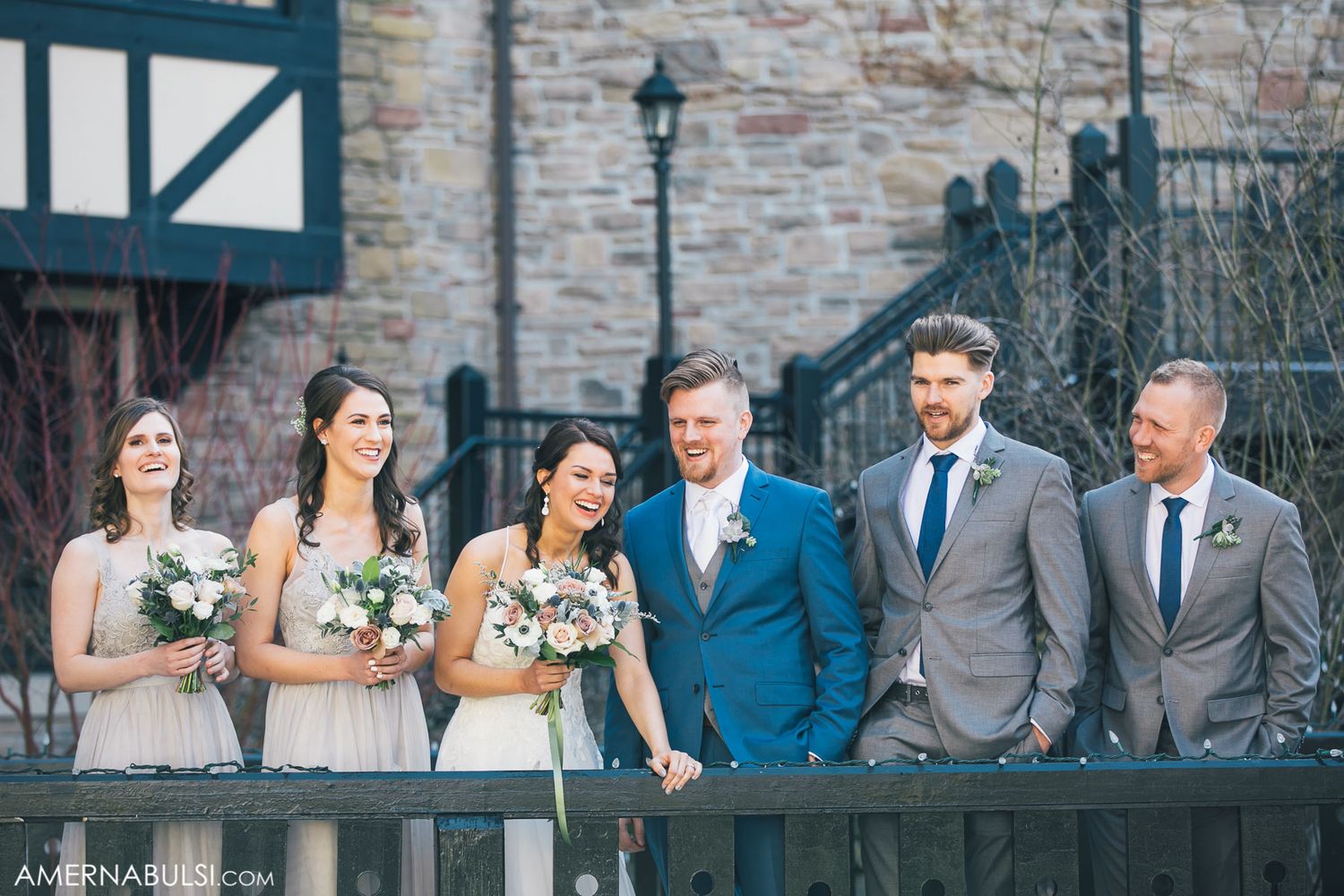 Wedding party in blue and gray attire posing against stone wall outdoors.