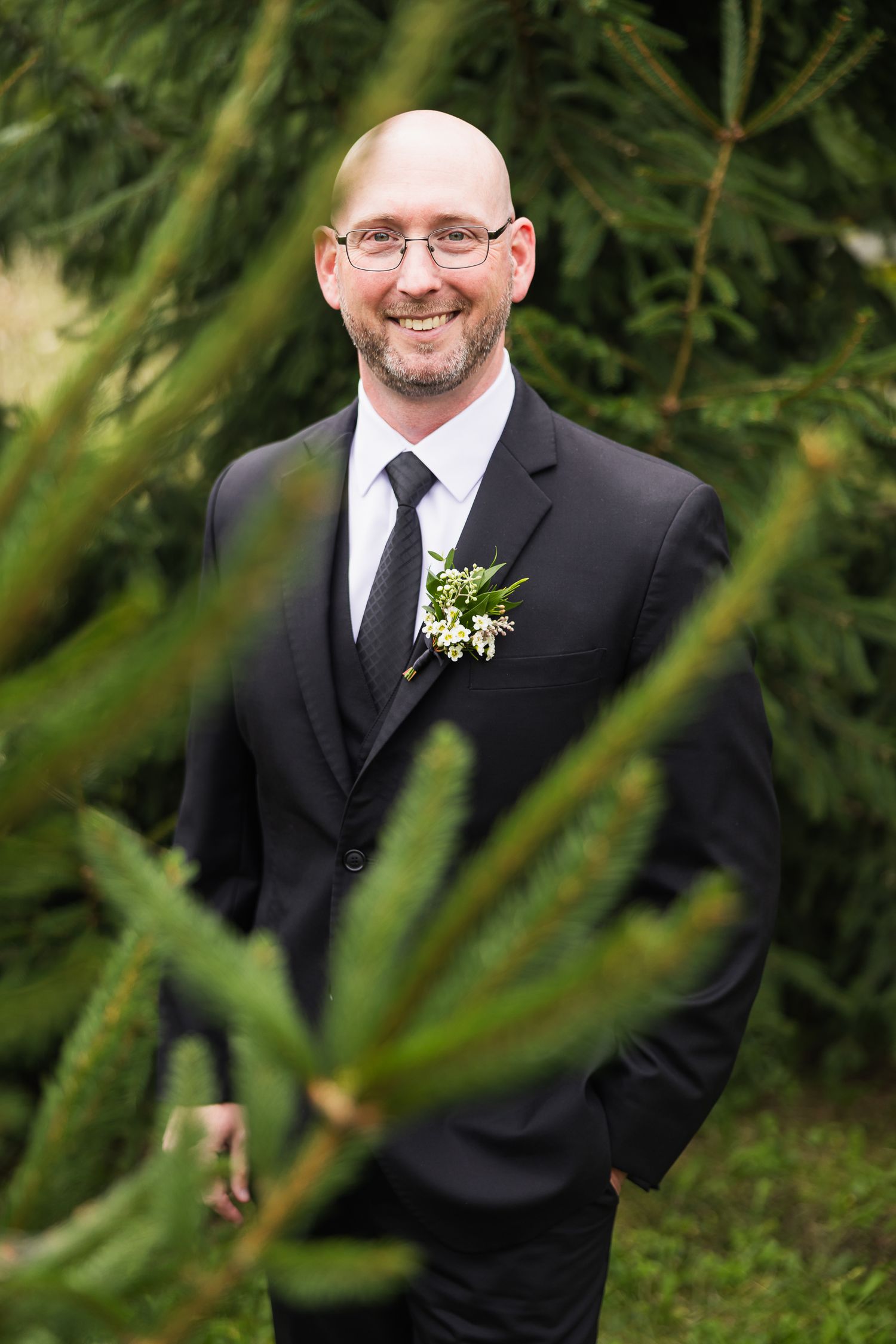 Wedding photos of someone wearing a black suit and boutonniere posing against an evergreen background in natural light.