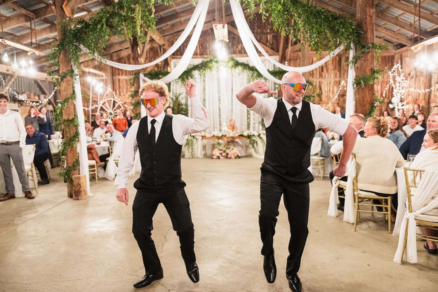 Line of groomsmen in black suits walking in sync down a rustic barn aisle decorated with greenery garlands.