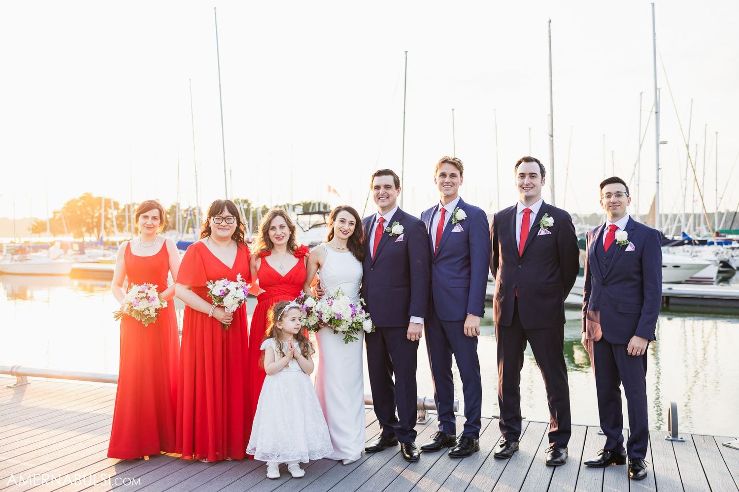 Wedding party in red and navy poses on wooden dock with marina backdrop.