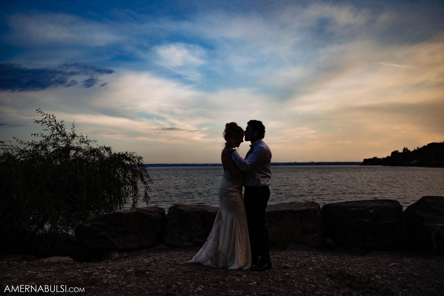Newlyweds embrace in silhouette against dramatic sunset over water.