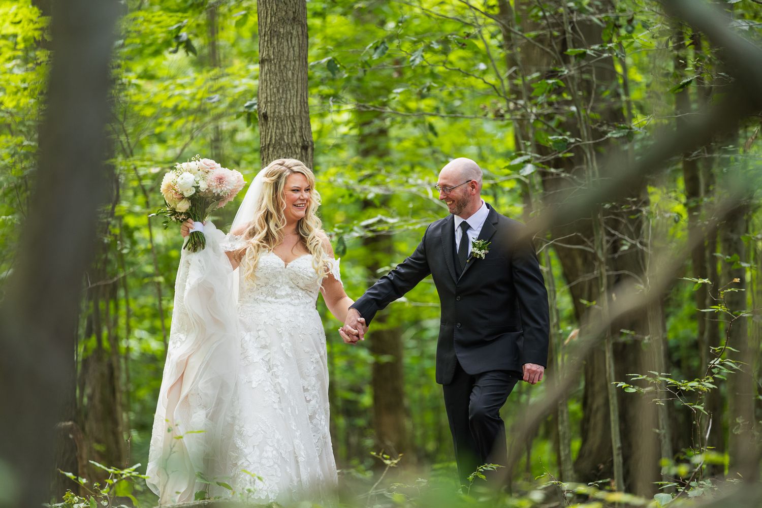 A joyful wedding couple walks hand in hand through a lush green forest setting during their special day.