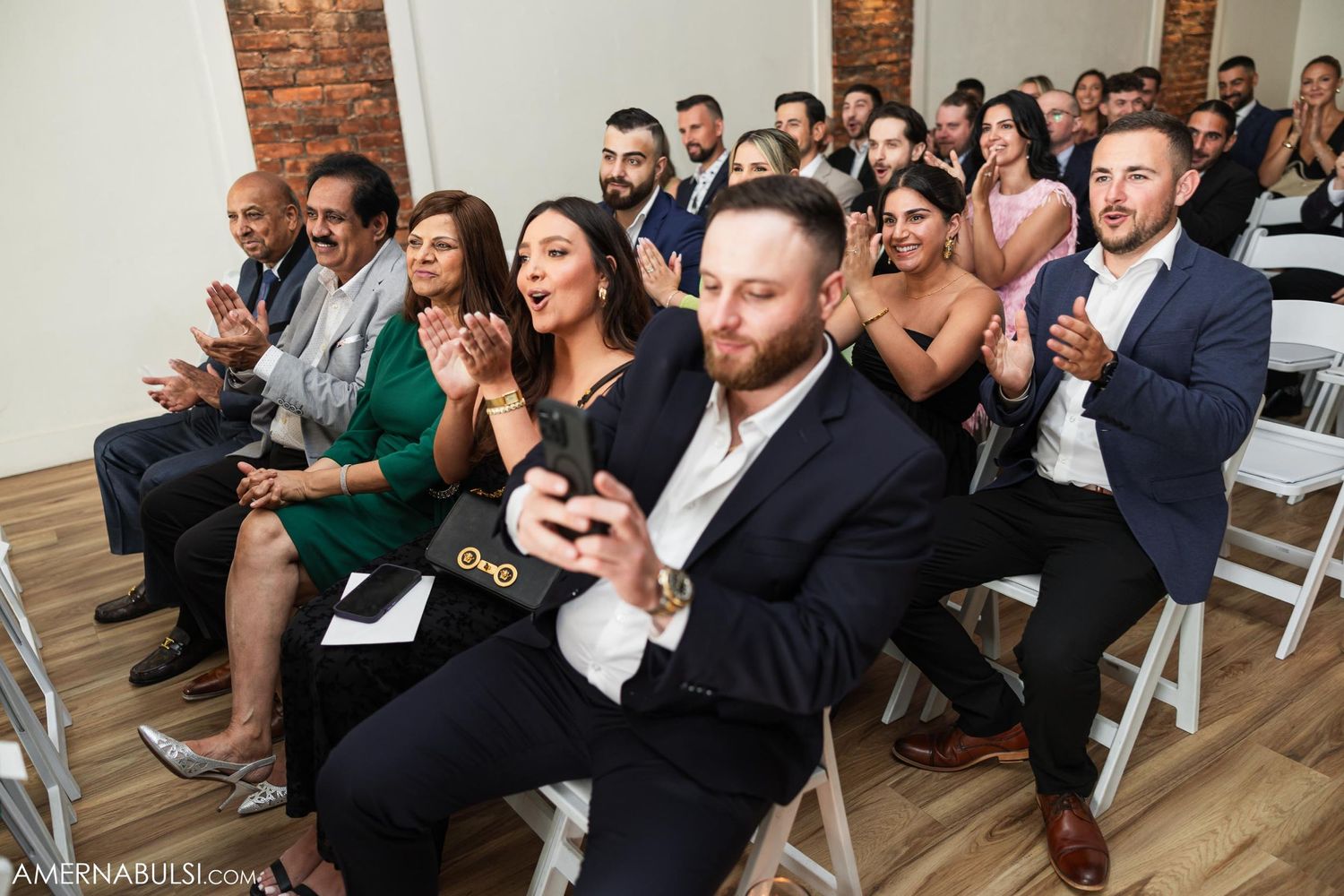 Wedding guests seated in white chairs at indoor ceremony venue with exposed brick walls.