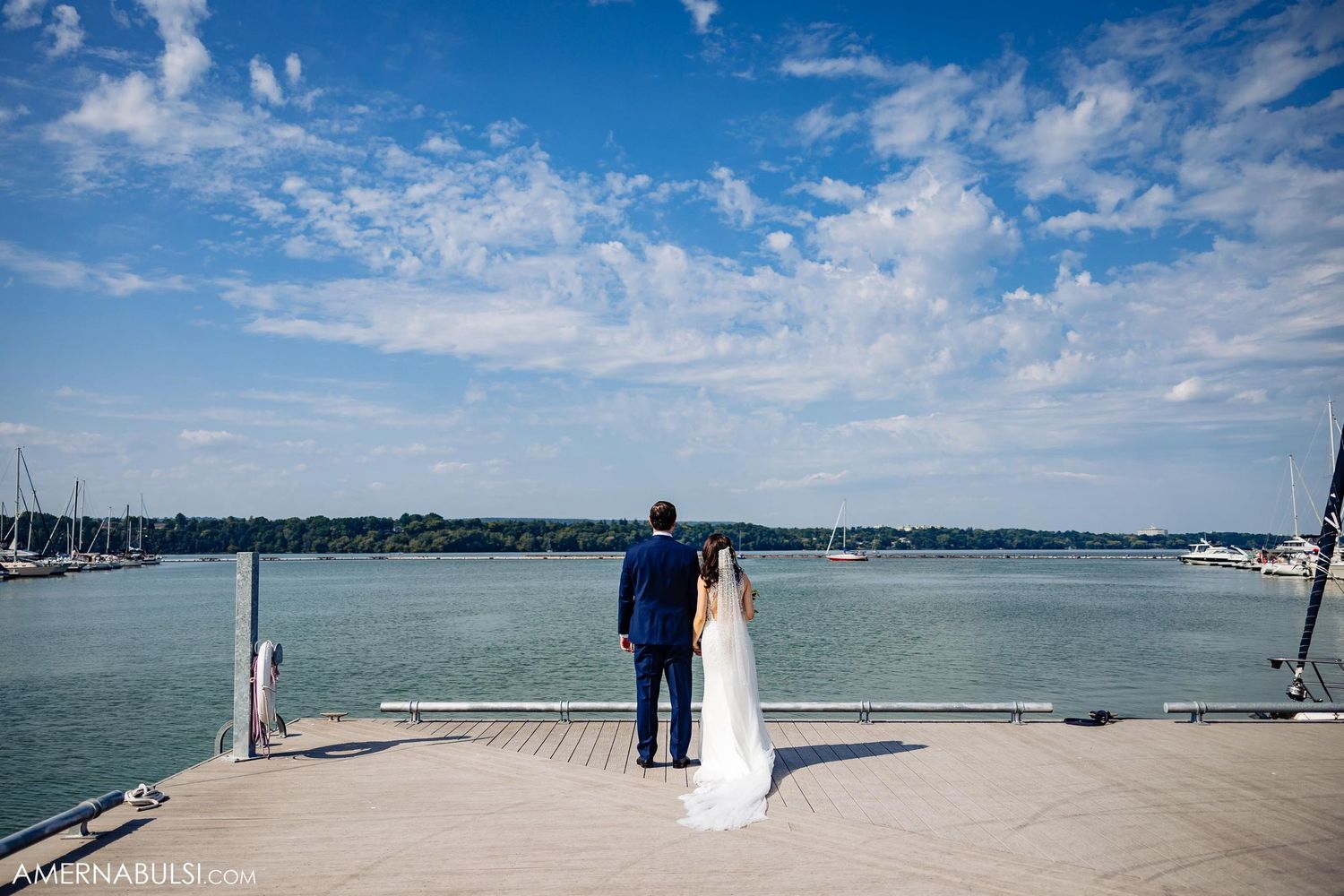 A couple stands together at the edge of a marina dock overlooking the water on their wedding day.
