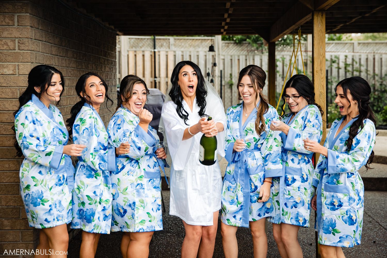 Bridesmaids in matching blue and white floral robes laughing together against brick wall.