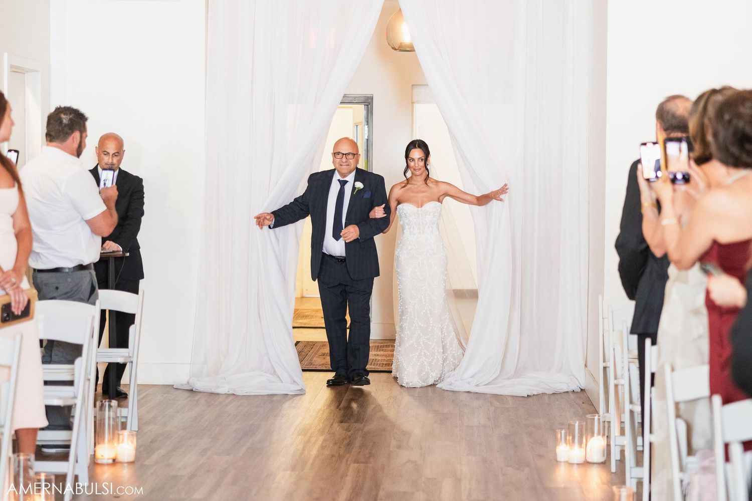 Father and bride make grand entrance down aisle lined with candles in industrial venue.