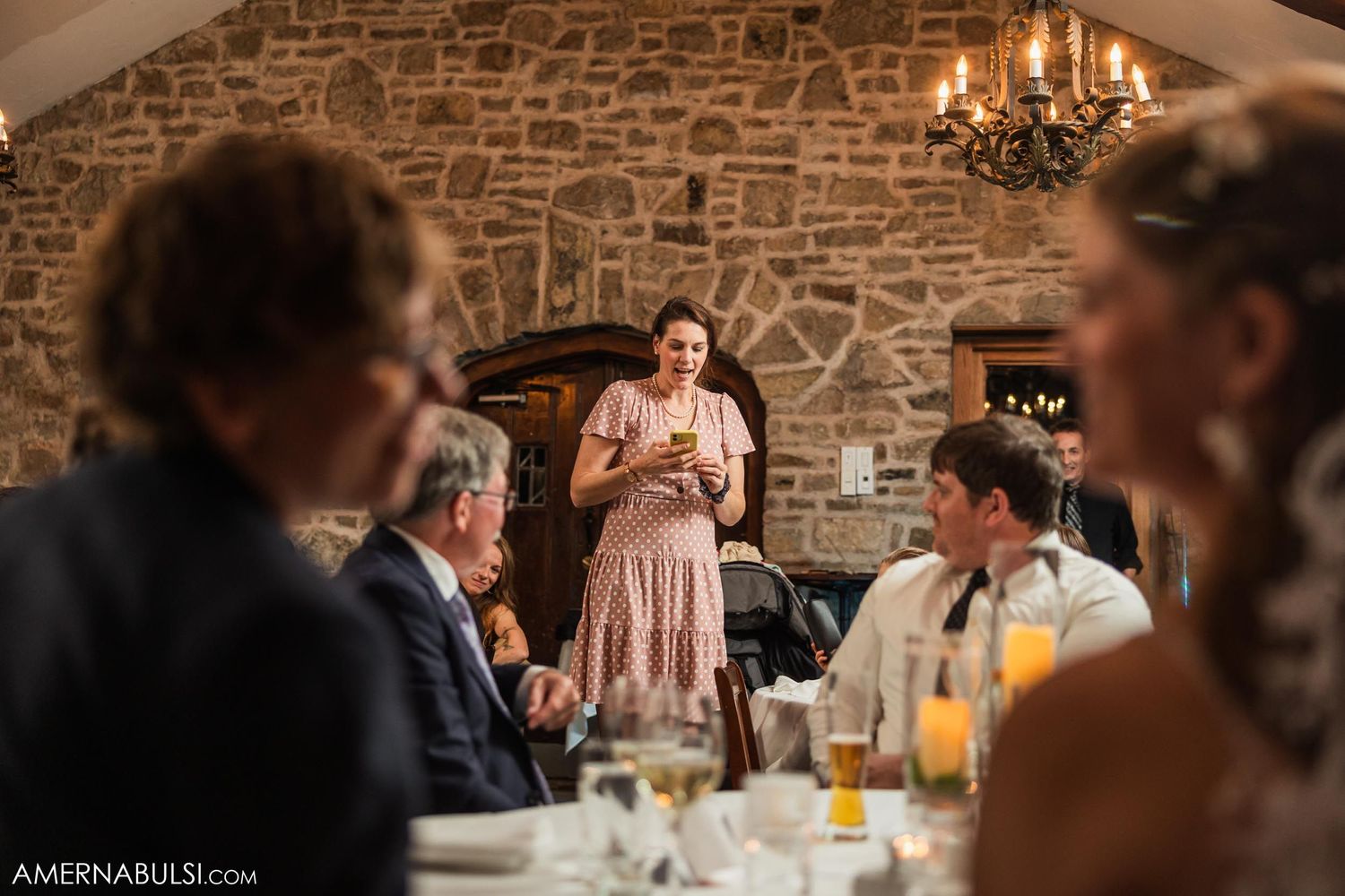 A guest delivers a wedding toast as others listen at tables in a stone-walled venue.