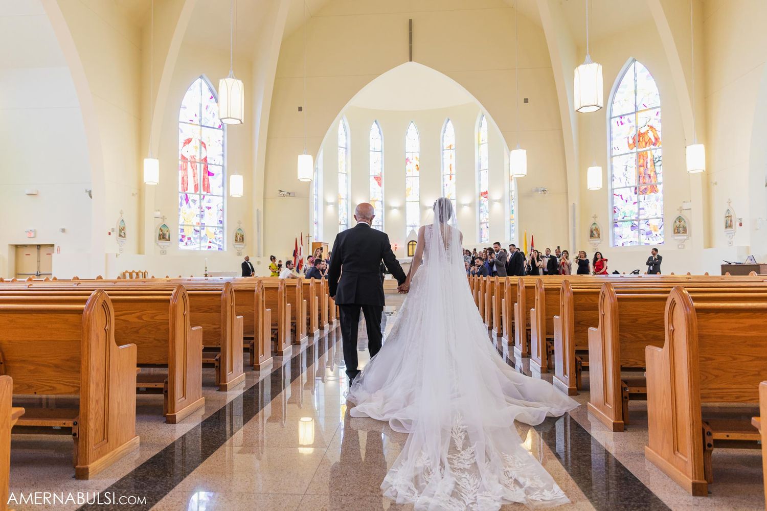 Bride and father walking down church aisle with long flowing veil trailing behind.
