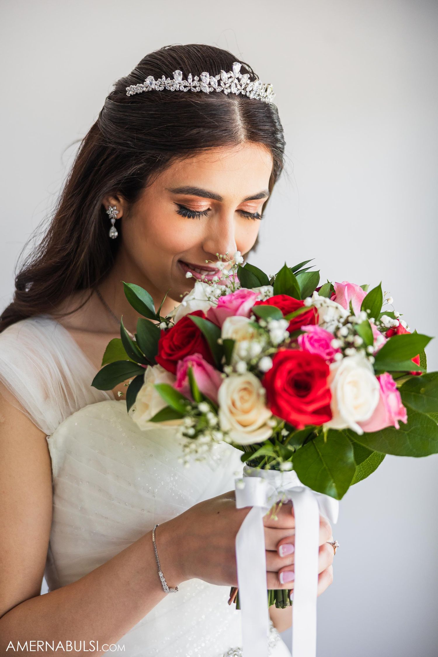 A bride in white holds a vibrant bouquet of pink and red roses while wearing a delicate tiara.