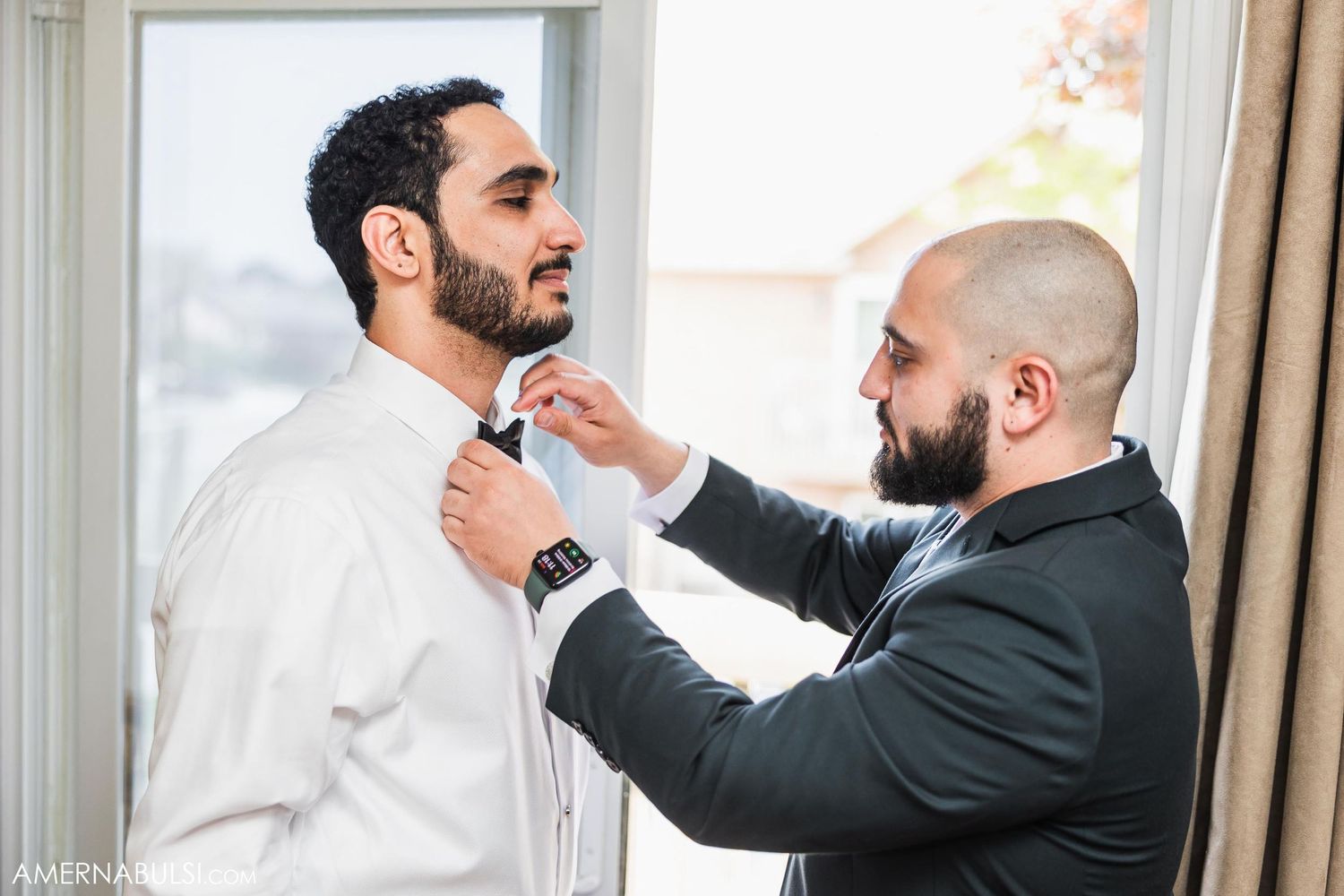 Two people prepare for a formal event as one helps adjust the other's bowtie.