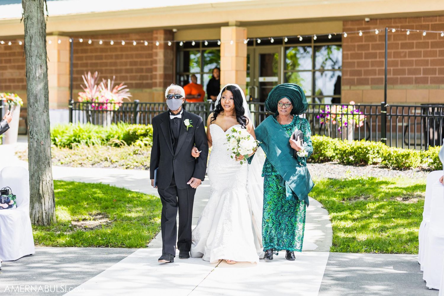 Wedding party members walk down outdoor path with string lights and brick building backdrop.