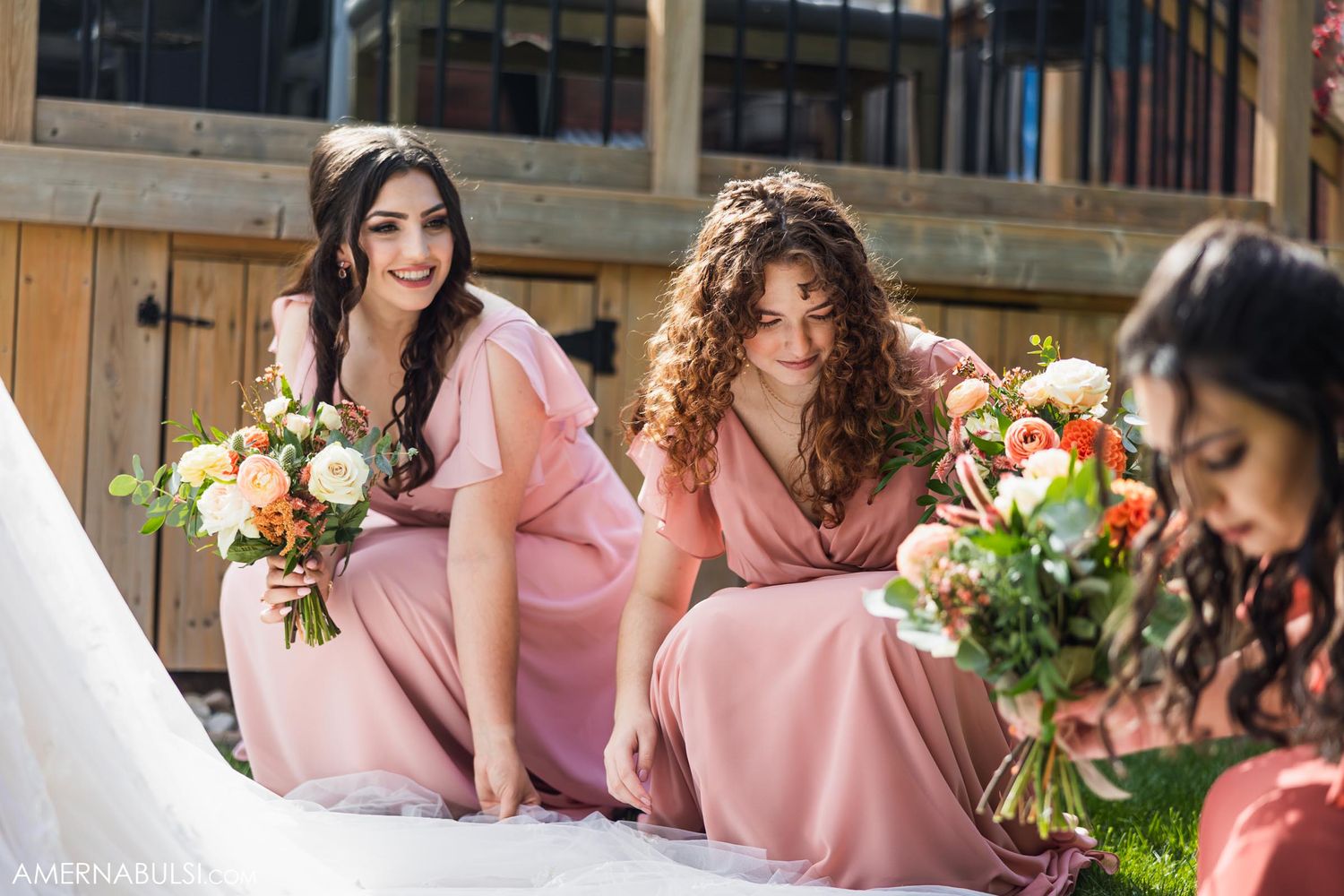 Bridesmaids in pink dresses relaxing on outdoor deck with colorful bouquets.