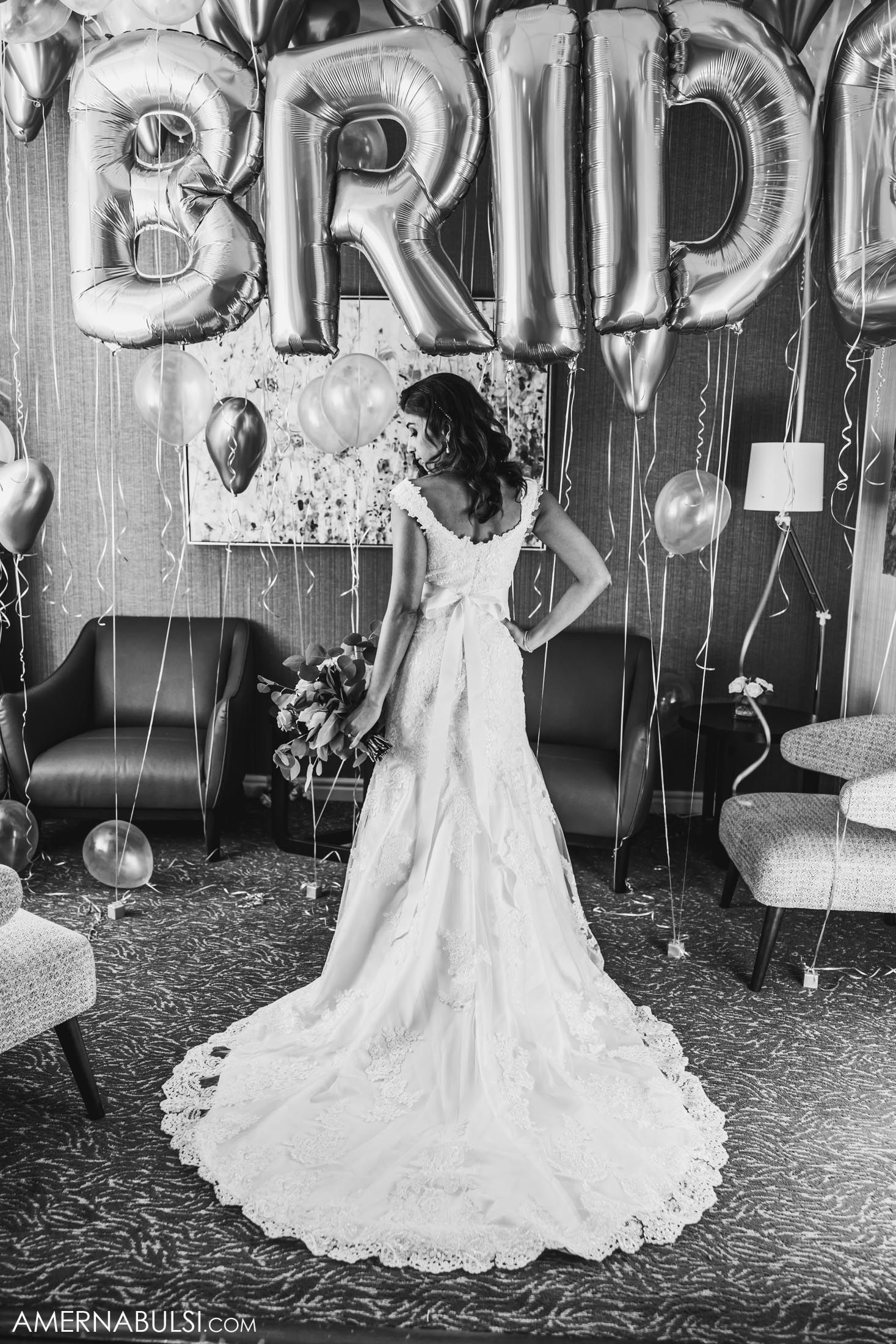 Elegant wedding dress with lace train shown from behind against silver BRIDE balloon backdrop and balloons.