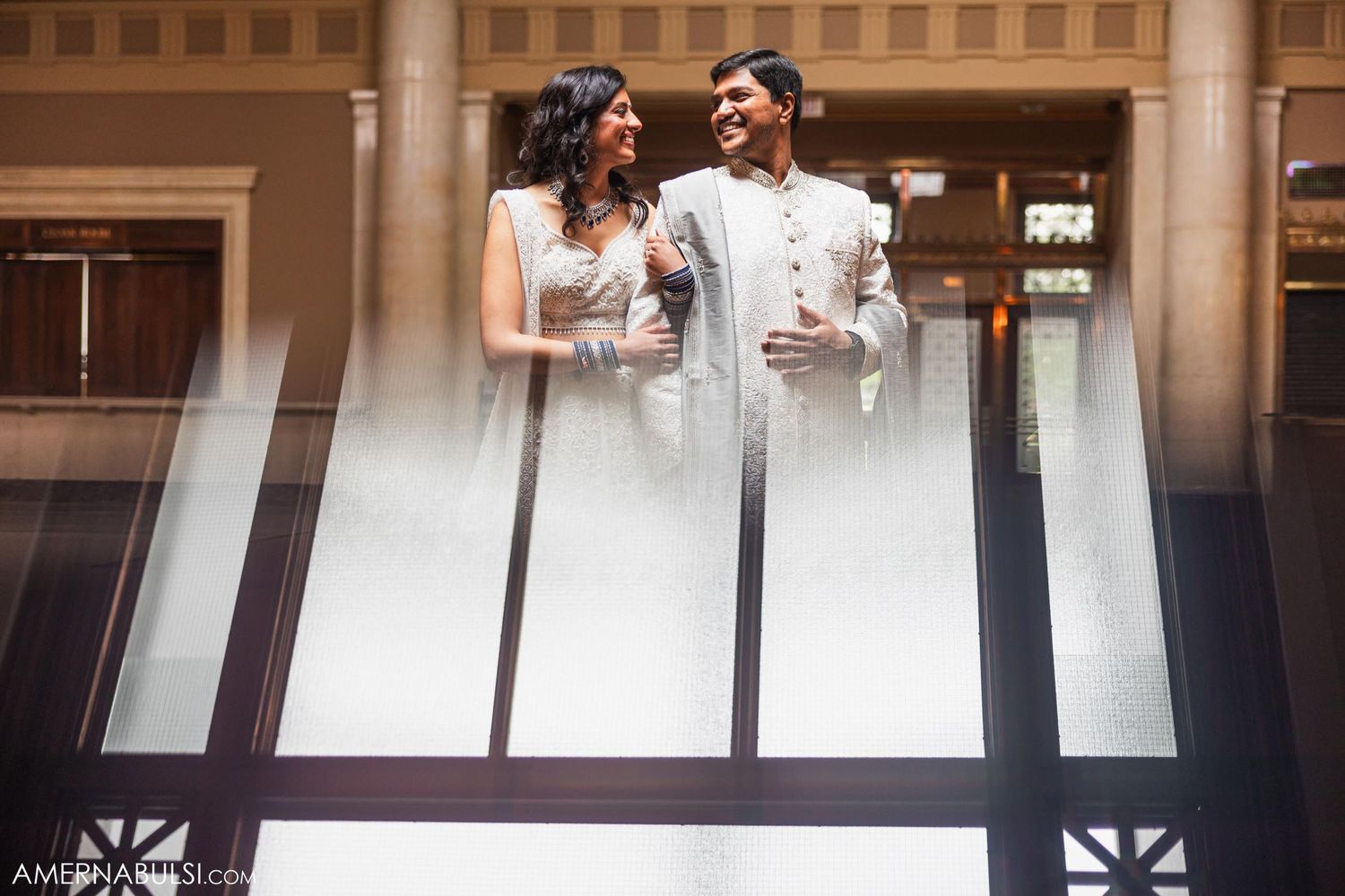 Couple in modern Indian wedding outfits pose by architectural elements with dramatic lighting.