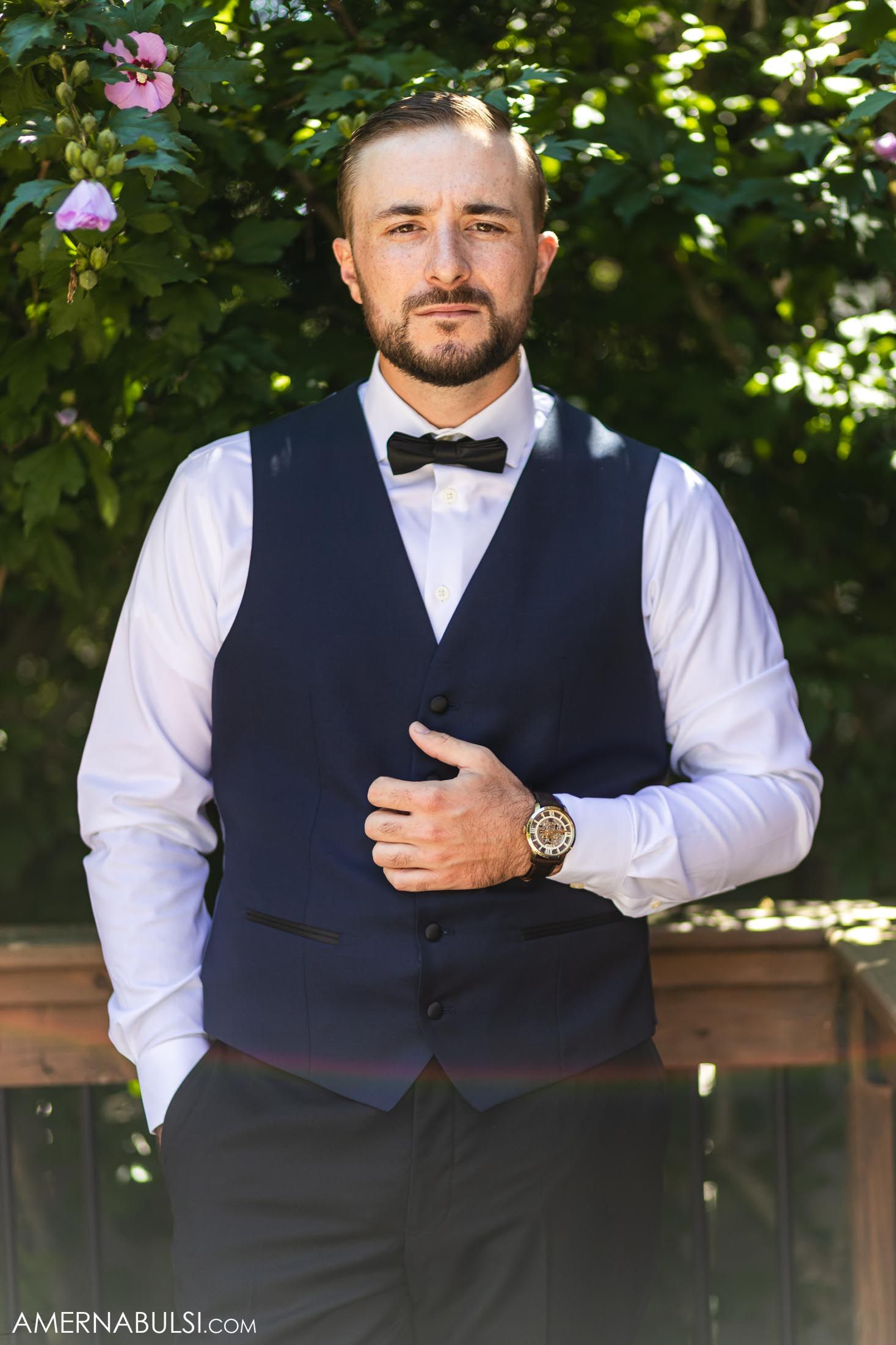 A person wearing a formal navy vest and bowtie stands in front of flowering bushes.