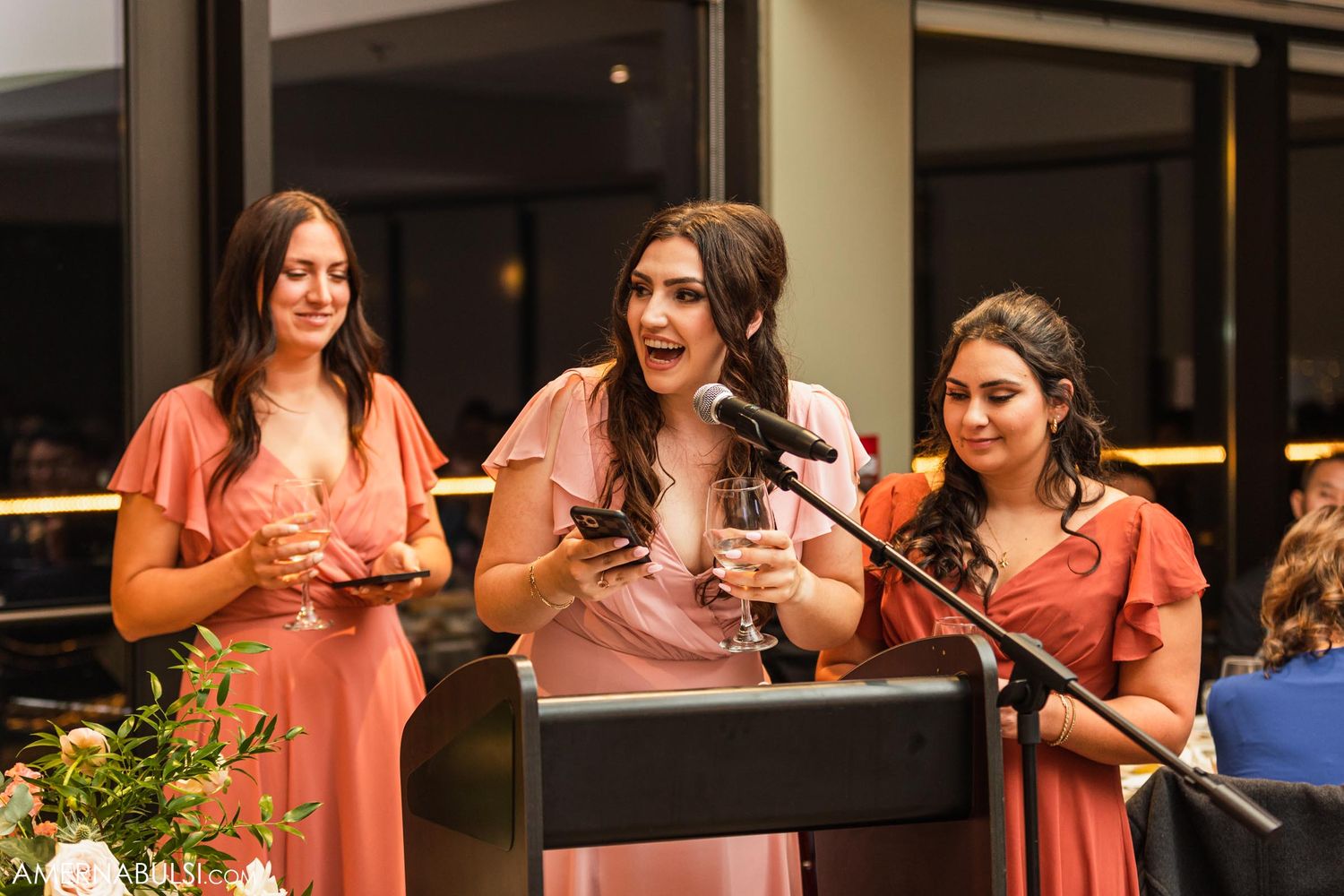 Three bridesmaids in coral-colored dresses deliver speeches at a wedding reception podium.
