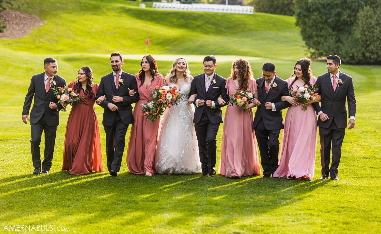 Wedding party in pink dresses and black suits poses together on sunny golf course.