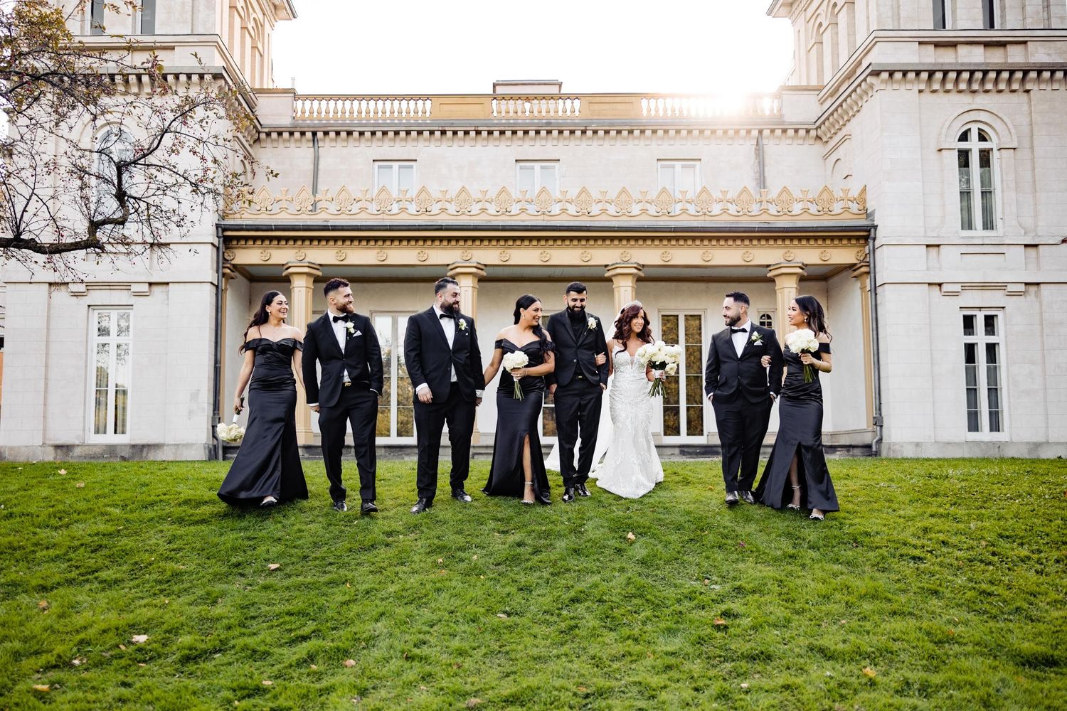 Wedding party stands casually on lawn in front of elegant white mansion.