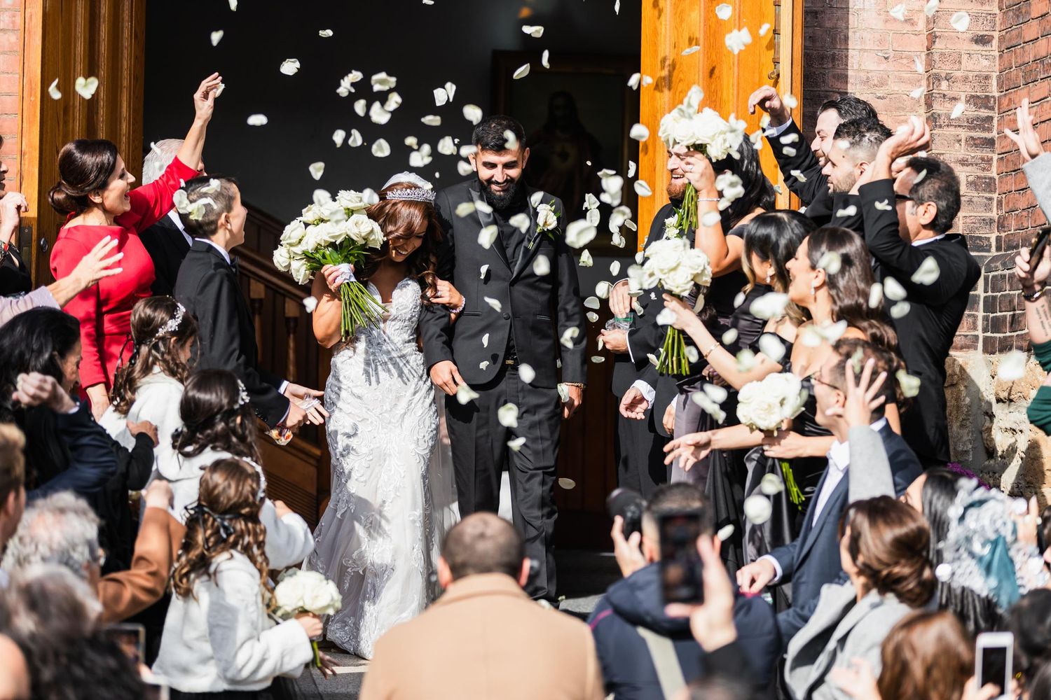 Wedding party exits church as guests shower them with white flower petals.