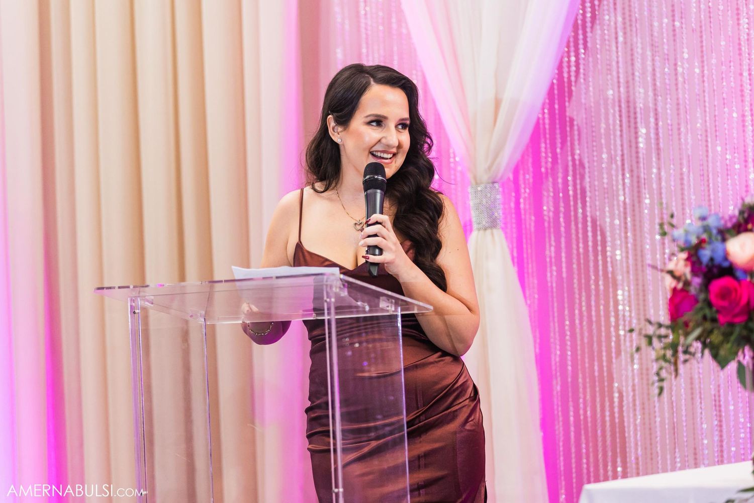 A speaker stands at a clear acrylic podium against a pink and cream draped backdrop with floral arrangements.