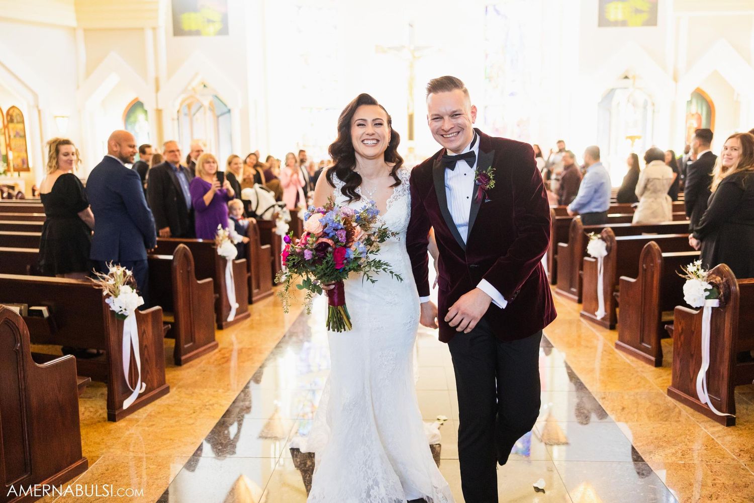 Couple walks down church aisle after ceremony with wooden pews and stained glass windows.