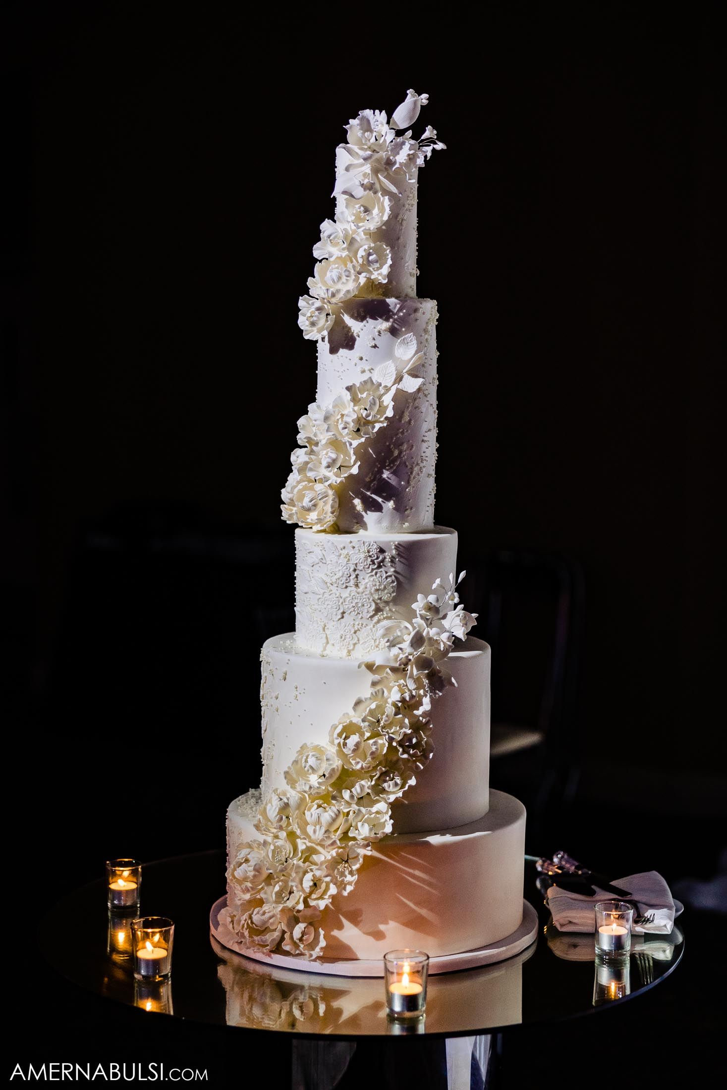 Five-tier white wedding cake adorned with cascading sugar flowers and illuminated by candlelight.