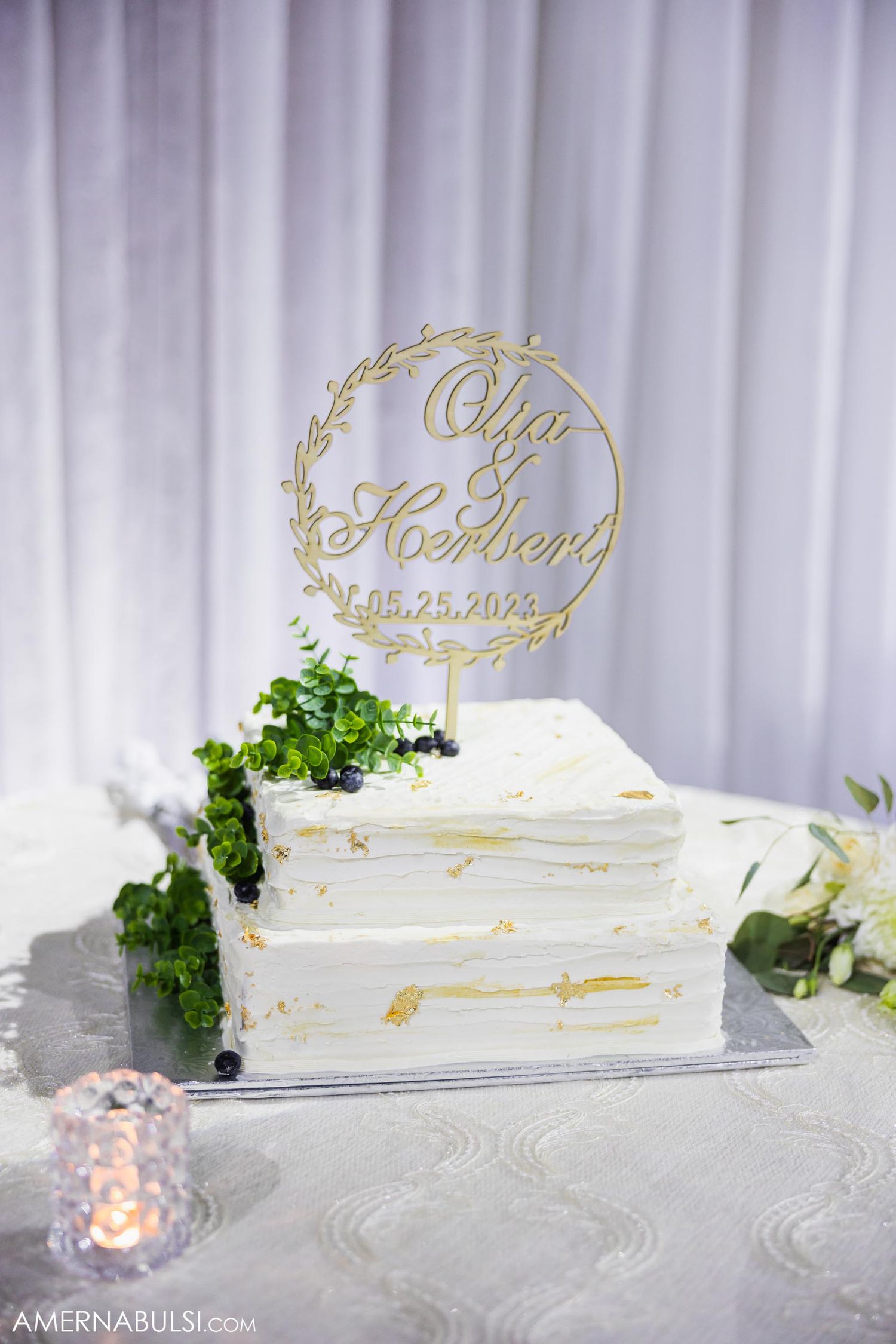 Elegant white wedding cake with gold decorative topper displaying names and date against a white backdrop.