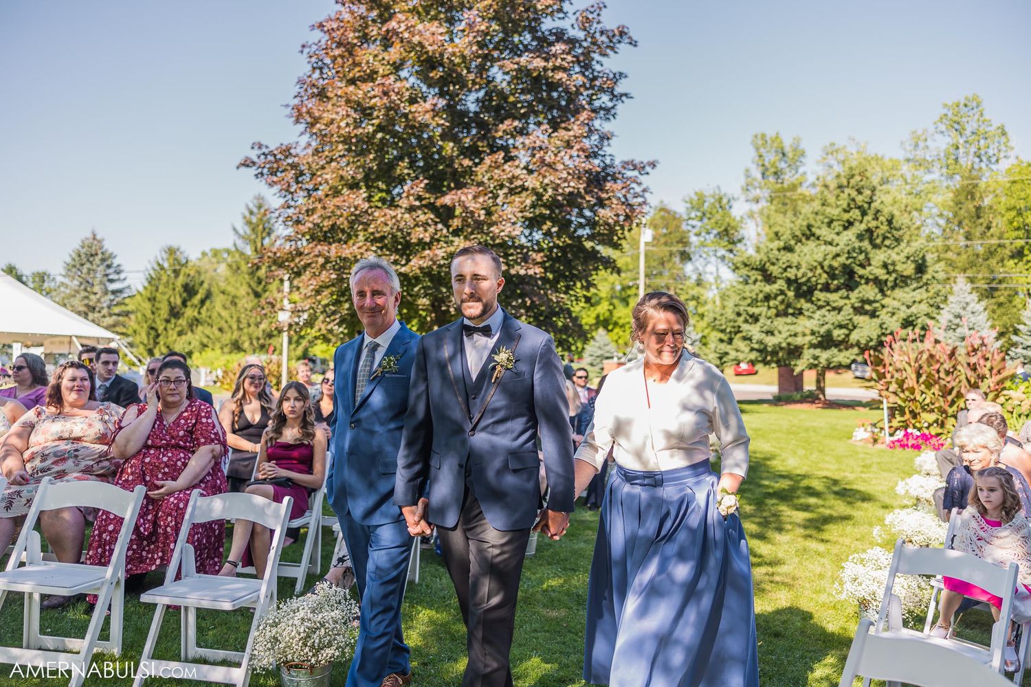 Outdoor wedding ceremony setup with white chairs on green lawn under trees.