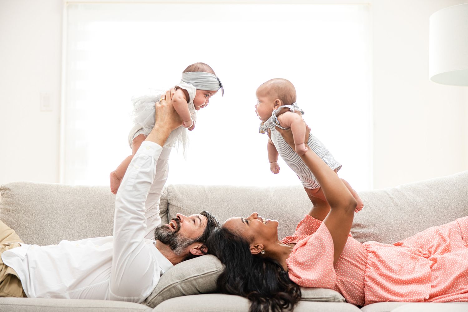 Two people lie on a white couch while playfully lifting babies overhead in a bright, airy room.