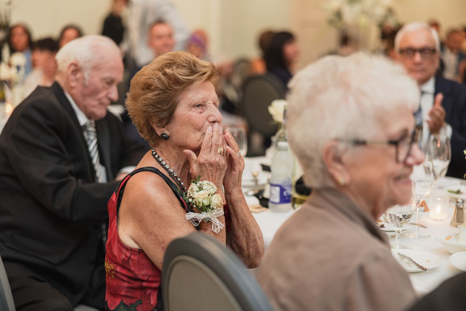 Elderly woman in red dress with corsage gestures emotionally at formal banquet dinner event.