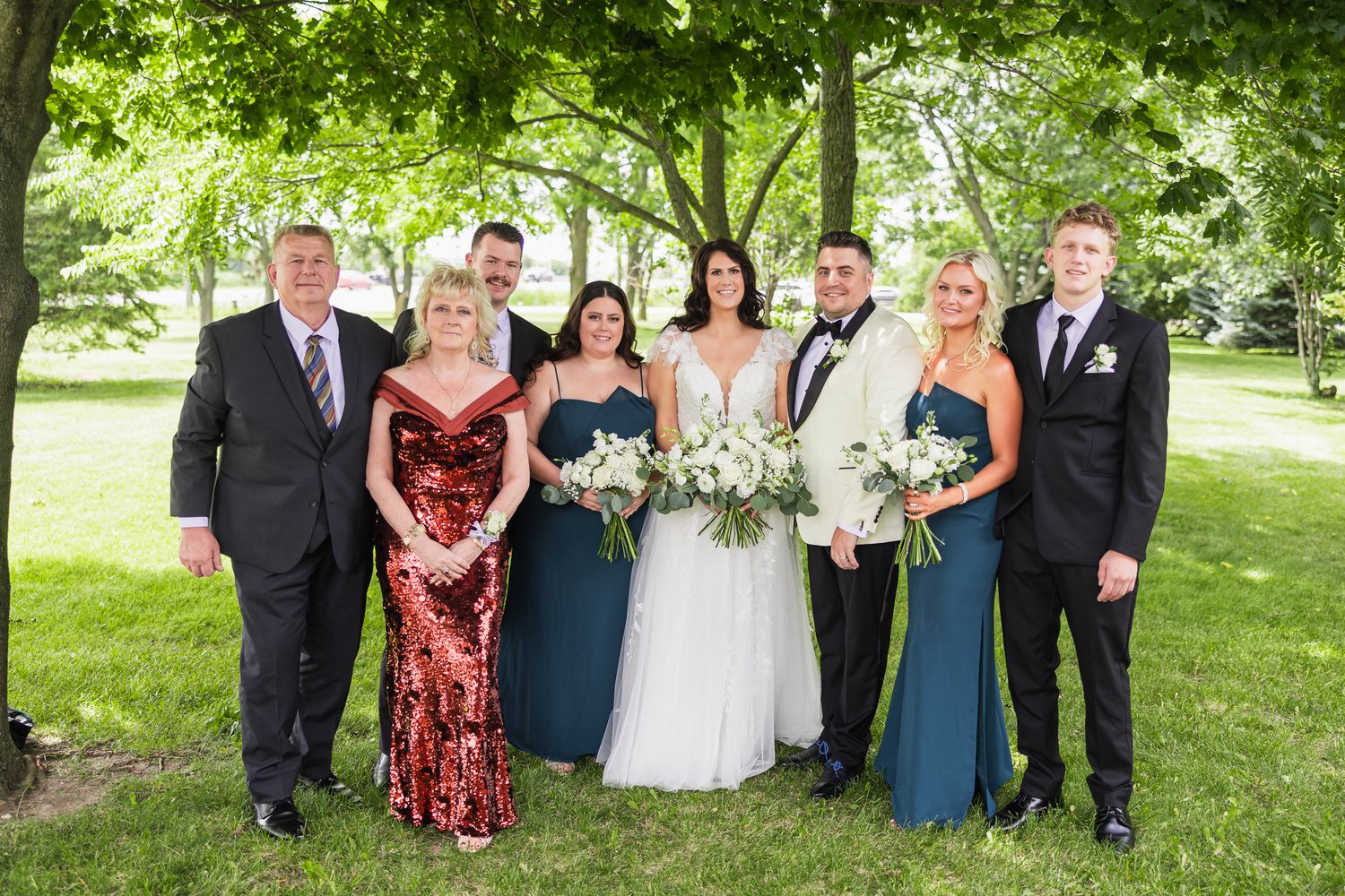 Wedding family group portrait outdoors with bride in white gown and groom in ivory tuxedo on green lawn.