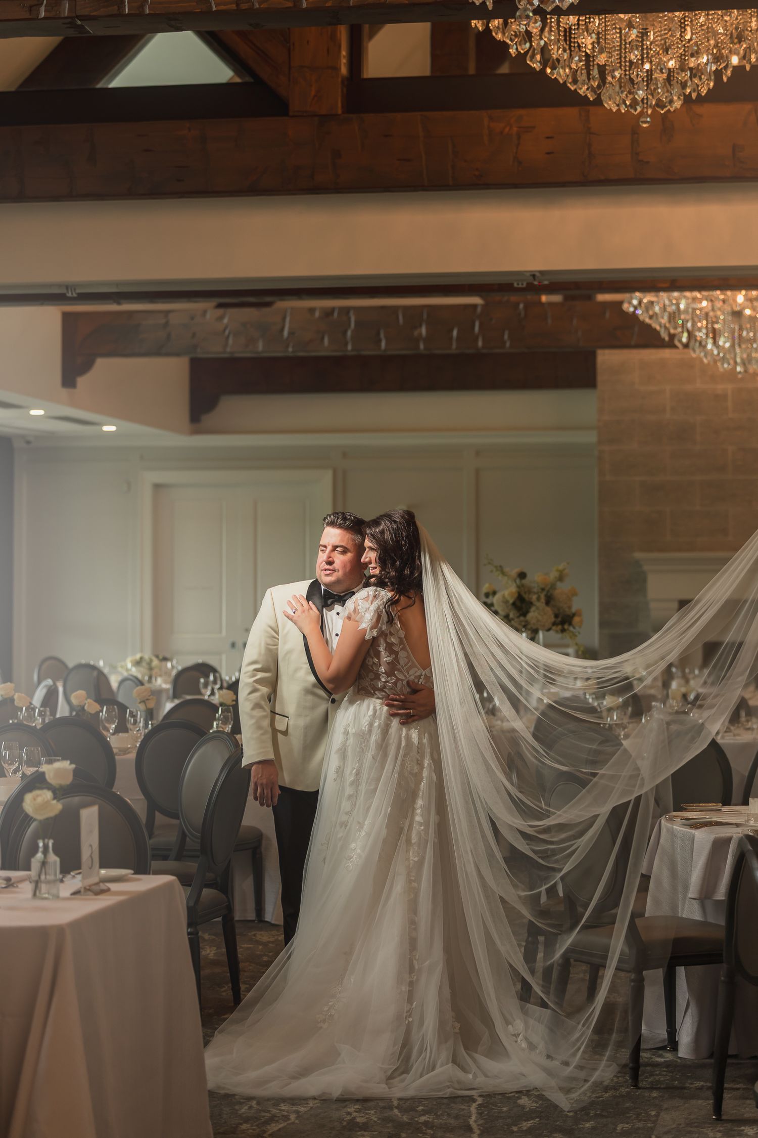 Bride and groom share romantic embrace in elegant reception hall with crystal chandeliers and flowing veil.