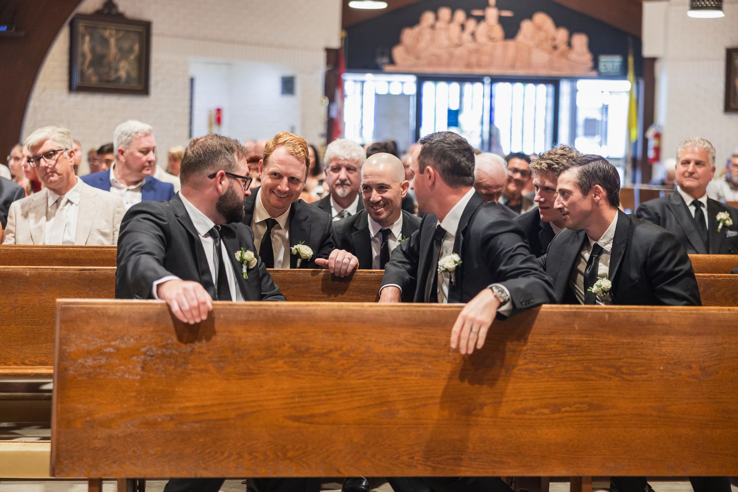 Groomsmen in dark suits with boutonnieres share a laugh together in church pews before a wedding ceremony.