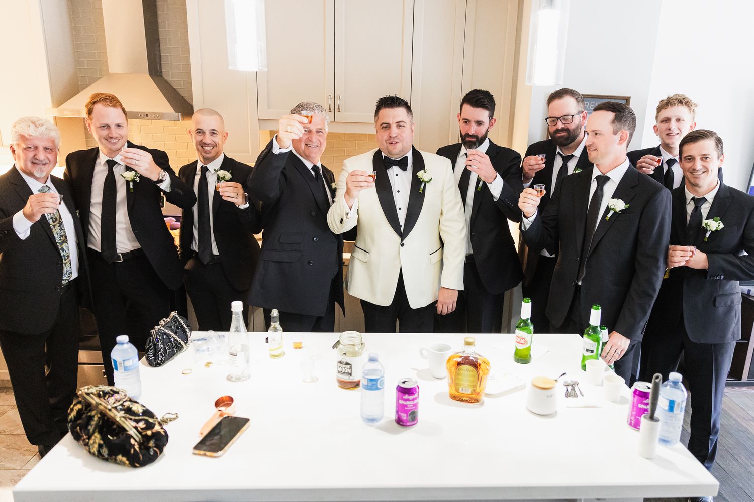 Groom in white tuxedo jacket raises a toast with groomsmen in black suits at a wedding reception table.