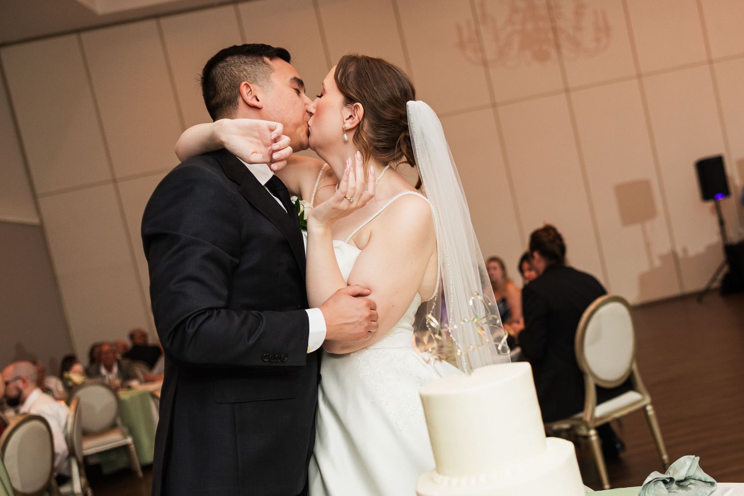 Newlyweds share first kiss beside white wedding cake at reception.