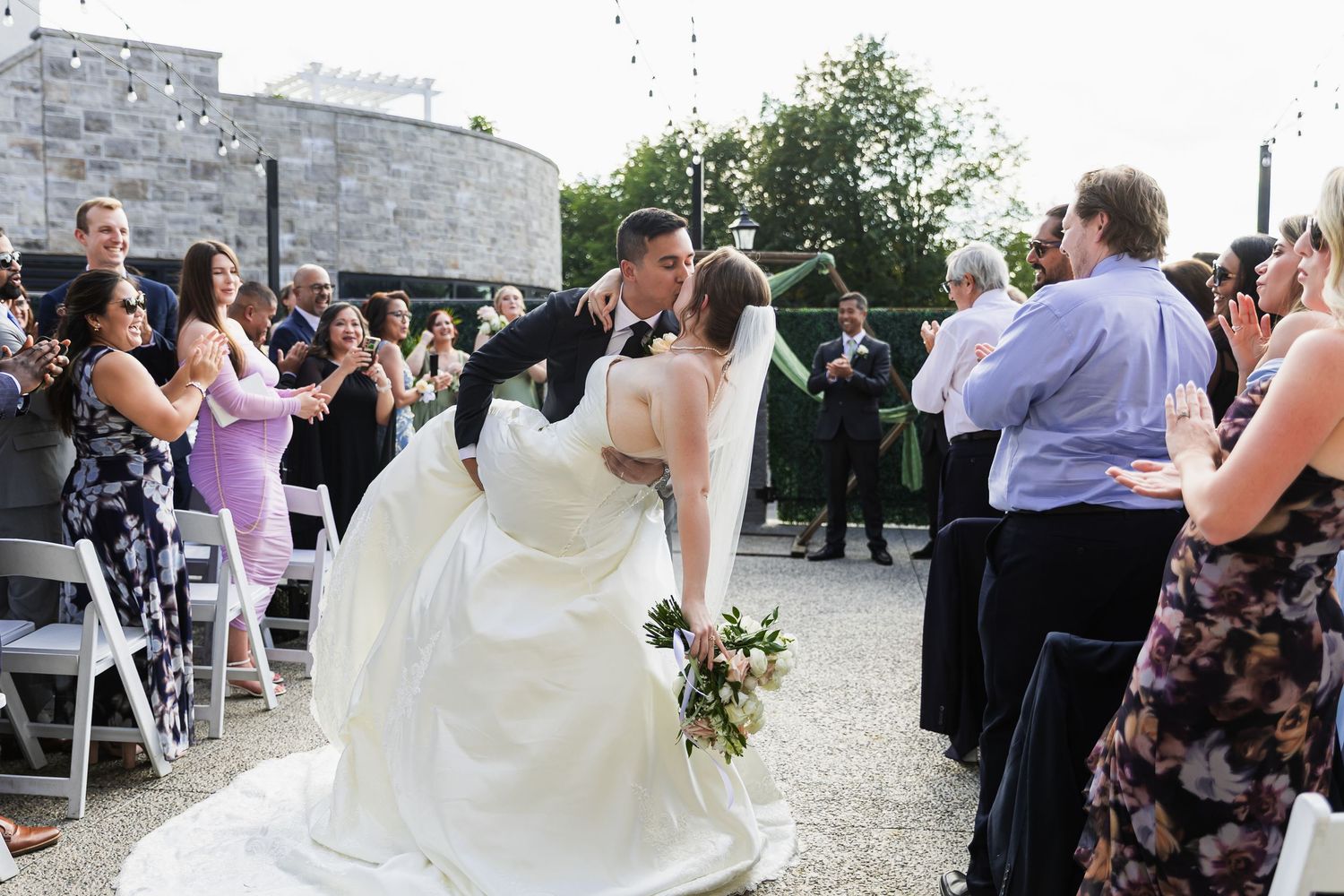 Groom dips bride for a kiss during outdoor wedding recessional as guests cheer and celebrate.